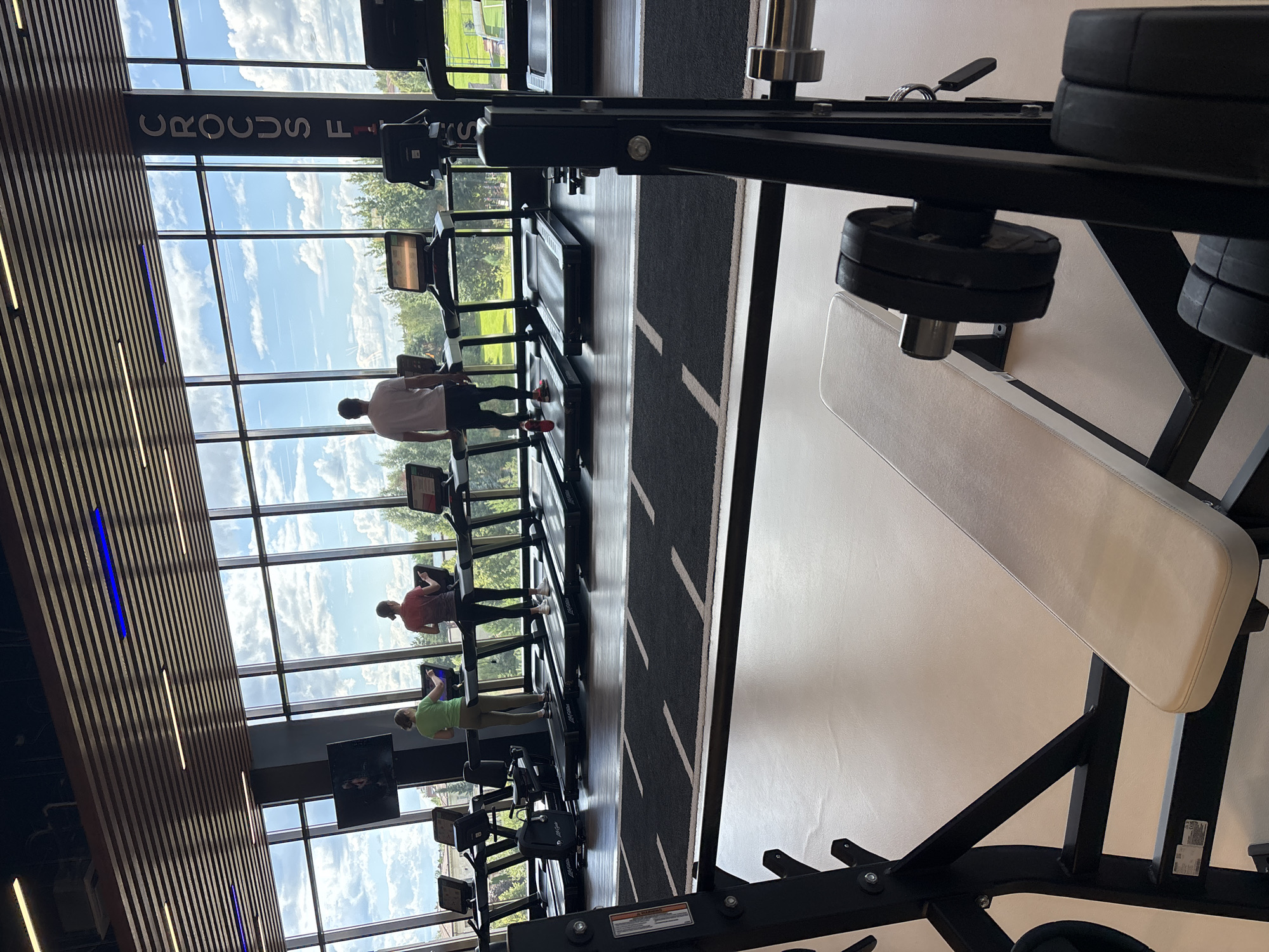 The image depicts the interior of a modern gym, likely located in Monolit, Russia, as suggested by the vertical text "CROCS F" visible on a pillar. The gym features a row of treadmills positioned in front of large windows offering a view of a treed landscape under a cloudy sky. Three individuals are seen using the treadmills. In the foreground, a weight bench with weights attached to a barbell is visible. The gym appears to have a clean and organized aesthetic with wooden ceiling panels and bright lighting.
