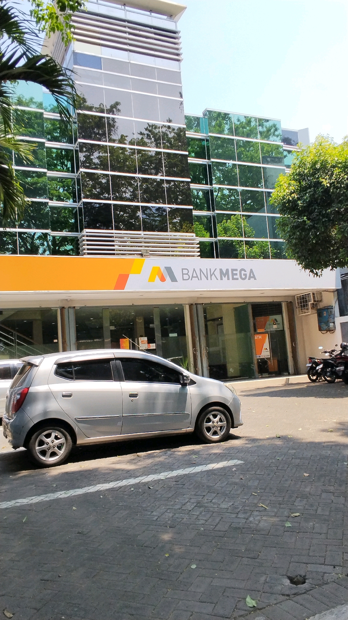 The image shows the exterior of a Bank Mega branch in Mojokerto, Indonesia. A silver hatchback car is parked in front of the bank. The bank building features a modern glass facade with reflections of trees and sky. The Bank Mega logo is prominently displayed above the entrance. In the background, two motorcycles are parked on the side of the road. The area appears to be a commercial district with paved sidewalks and a street. The lighting suggests it is daytime, likely a sunny day.