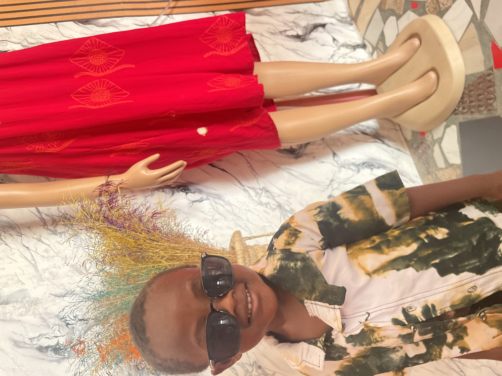The scene depicts a person wearing sunglasses and a shirt with a mottled green and beige pattern, lying next to a mannequin. The mannequin, dressed in a red dress featuring decorative sunburst patterns, has its legs displayed on a wooden stand. Beside the person's head is a bundle of dyed straw in various colors. The setting appears to be indoors, possibly in Badagry, Nigeria, based on the prompt, with a marble-patterned surface beneath the person and mannequin. A piece of striped wood paneling is visible in the background above the mannequin. The orientation of the photograph is sideways.
