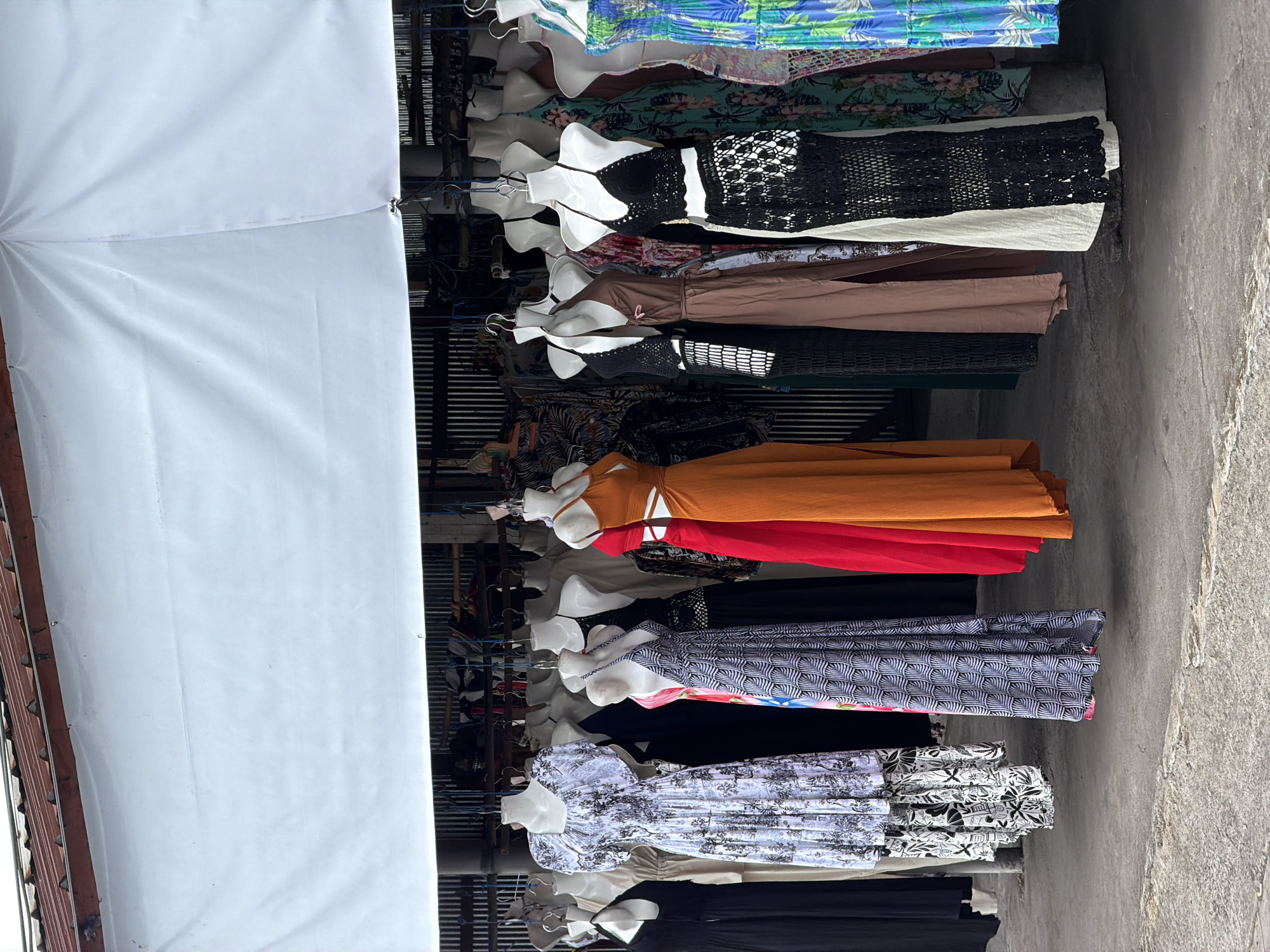 This image depicts a clothing stall in Kintamani, Indonesia, specializing in flowing dresses and beachwear. The stall features several mannequins adorned with various styles of dresses, ranging from long, patterned maxi dresses to shorter, brightly colored outfits. The clothing is displayed under a white tarp that provides shade, and behind the mannequins, a collection of clothes on hangers is visible, hinting at a wider selection. The ground is a rough, paved surface, and the background suggests a simple, open-air market setting with wooden structures and corrugated metal roofing. The scene is devoid of any people, focusing solely on the merchandise.