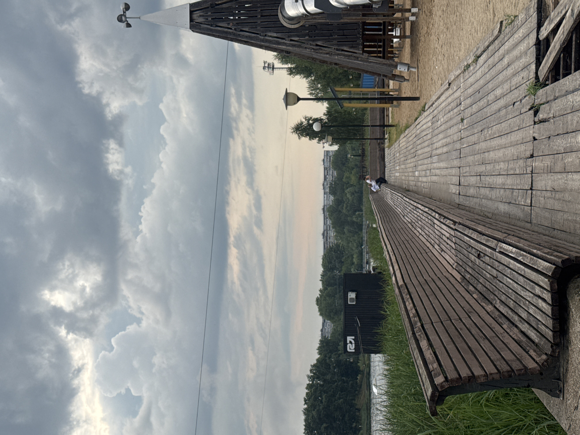 The image depicts a waterfront recreational area, possibly a park or a sports complex, with a wooden walkway and seating area. The scene is set against a backdrop of a cloudy sky, a body of water, and lush green trees lining the opposite bank.
In the foreground, a long, curved wooden bench runs along the edge of the walkway. The bench is constructed from numerous horizontal wooden slats, showing signs of weathering and age. The walkway itself is also made of wooden planks, some of which appear to be damaged or uneven.
To the right of the walkway, a large, conical wooden structure, likely part of a playground or an observation tower, dominates the scene. It features a dark wooden frame and a white spire at the top, topped with a weather vane. A large metal slide is partially visible, descending from the structure. The ground around this structure is sandy.
Further back, across the water, a row of dense trees forms a natural barrier, behind which buildings of varying heights are visible, suggesting an urban or suburban setting. On the left bank, a dark, rectangular structure with the logo "R2I" in white is situated near the water's edge. It appears to be a small building or a changing room, possibly for water sports. The water itself is calm, with some ripples visible.
In the mid-ground, two individuals are seated on the wooden bench. One person, wearing a white shirt, is looking down, while the other, also in white, is seated next to them. Several lampposts are strategically placed along the walkway, indicating a well-equipped public space.
The sky is filled with heavy, grey clouds, suggesting an overcast day, potentially with impending rain. The overall impression is that of a functional, somewhat rustic, outdoor leisure facility. Based on the "R2I" logo, which is associated with the R2I Wake Park in Nizhny Novgorod, Russia, the location is likely this wakeboarding and recreational complex.