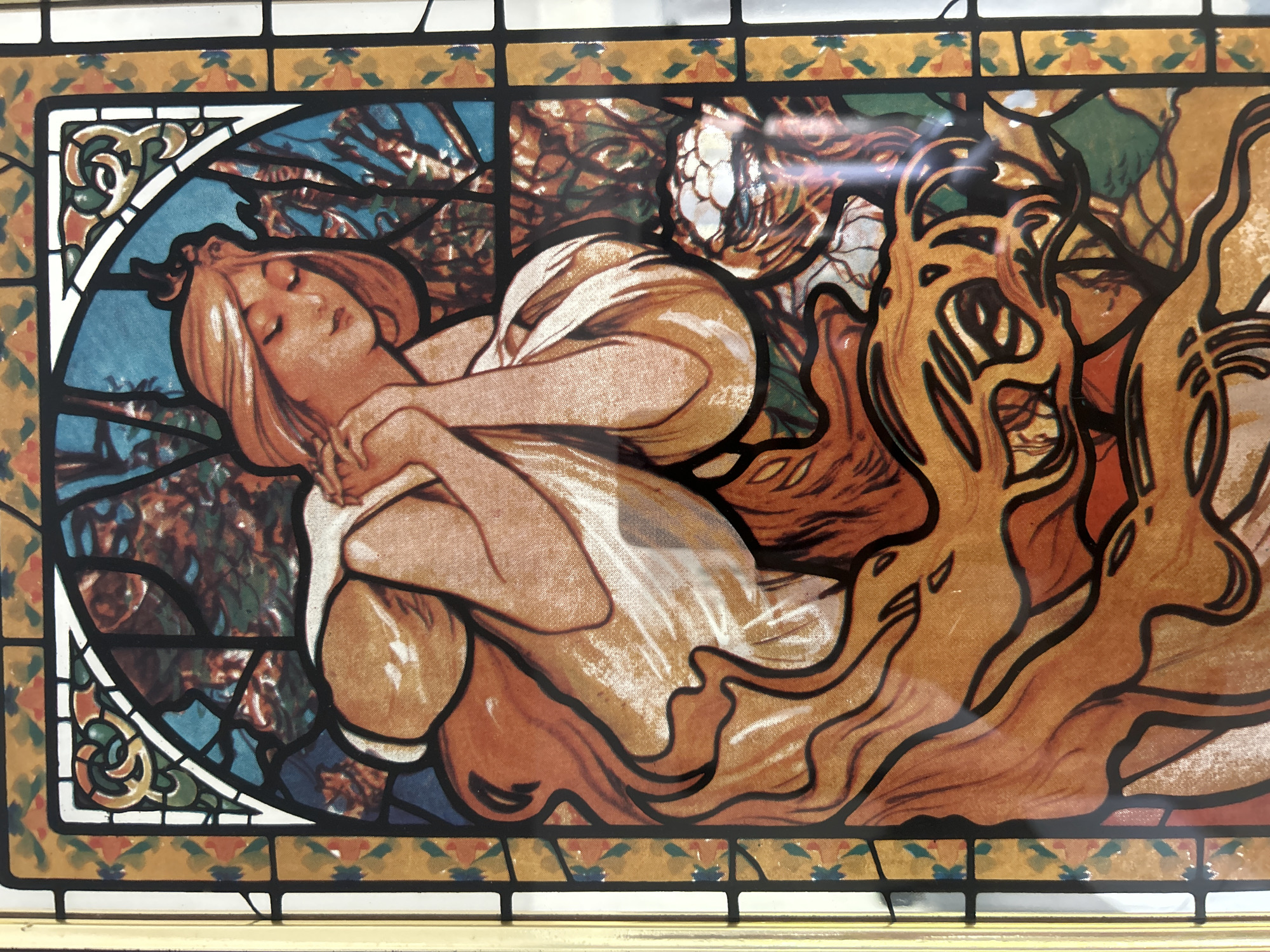 The image is a close-up of a framed stained-glass style artwork. The artwork depicts a reclining woman with fair skin and reddish-blonde hair, eyes closed, and arms crossed over her chest. She is draped in a loose, white robe with orange highlights and shadows. The background is a mix of blue and brown hues, resembling foliage. To the right of the woman, there are stylized, flame-like forms in shades of orange and brown. The frame is made of a repeating geometric pattern of square segments in black, brown, gold, and white, with colorful ornamental design.
There is no information available that indicates a location within the city of Câmara de Lobos, Portugal.