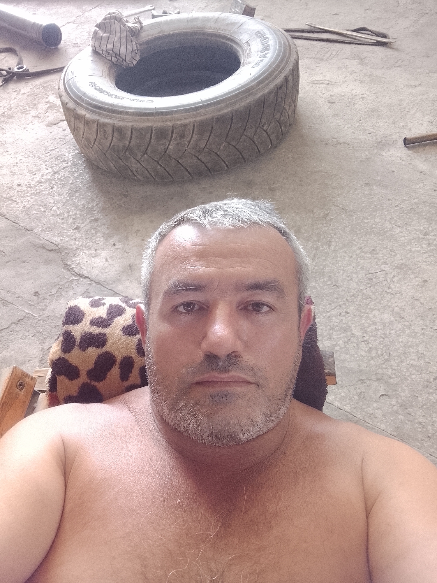 In Sabir Rakhimov, Uzbekistan, a man with graying hair and a beard is pictured from the chest up, seated. He is shirtless and resting his back against a patterned cushion. Behind him, a worn tire lies on a concrete surface, with a piece of striped cloth placed inside it. Various tools and metal objects are scattered on the concrete floor around the tire, suggesting a workshop or garage environment. The lighting is dim.
