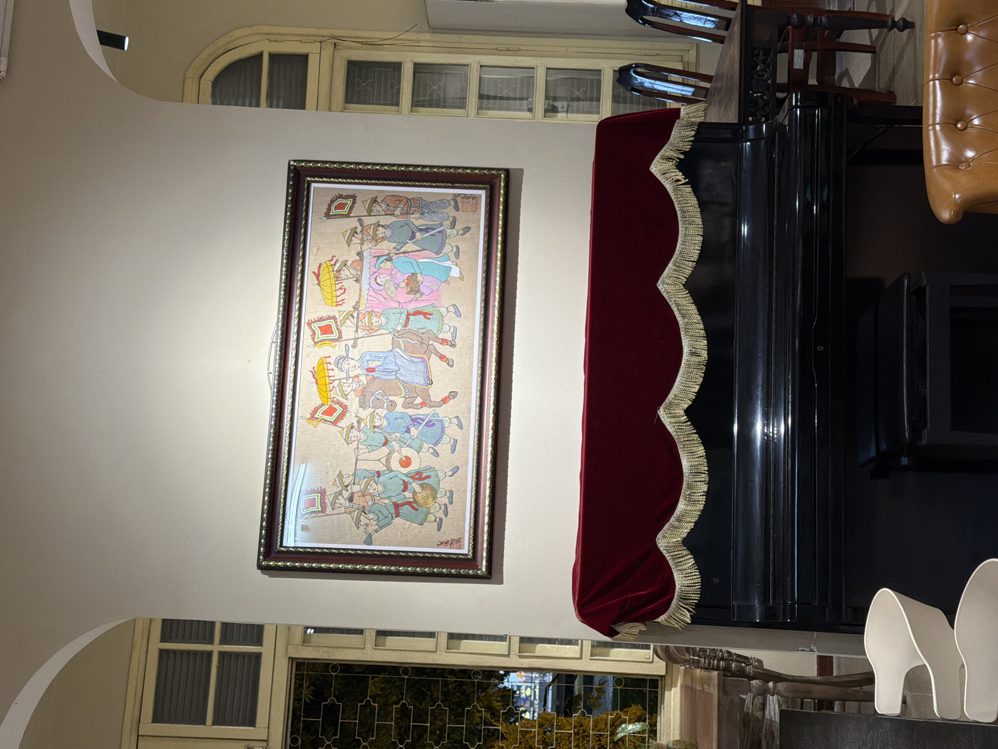 The image shows an indoor scene with a large framed painting of a procession displayed on a cream-colored wall. Below the painting is an upright piano covered with a deep red velvet cloth that has gold fringe. To the right of the piano is a dark wooden table with several wooden chairs. To the left of the piano, a portion of a wooden table is visible, along with a white chair. In the background, tall windows with wooden frames and panes are visible. The overall impression is of a dimly lit, perhaps antique or traditional, interior space.