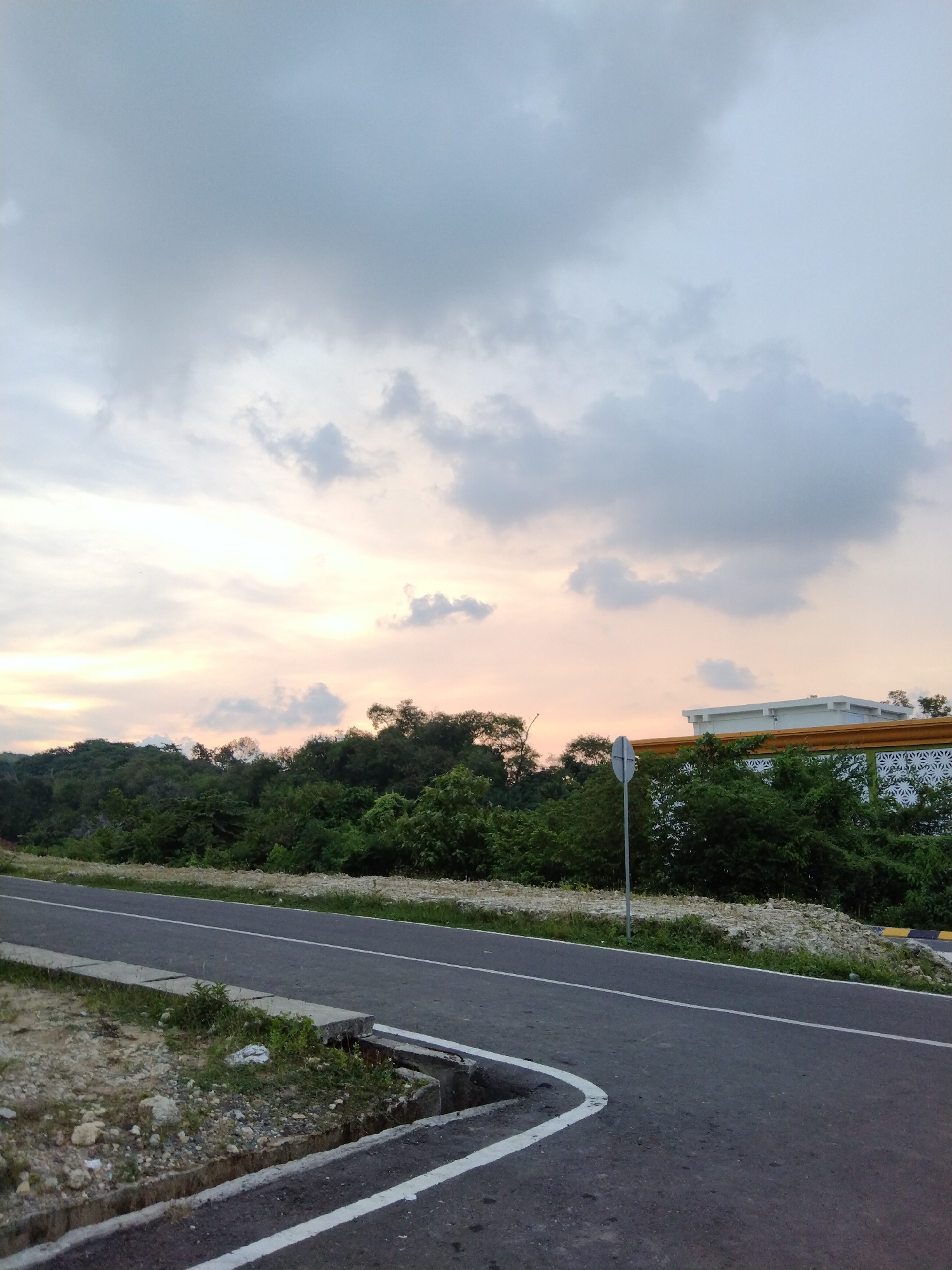 The image depicts a serene outdoor scene featuring a paved road, dense vegetation, and a cloudy sky. A two-lane asphalt road with clear white lines, one marking the edge and another serving as a dashed center line, extends from the foreground towards the background on the right. A concrete drainage ditch runs along the left side of the road, bordered by loose dirt, small rocks, and sparse green weeds.
To the right of the road, a thick band of lush, dark green trees and shrubs forms a verdant landscape, suggesting a natural or undeveloped area. Further in the background, partially visible above the tree line on the right, is a modern-looking building. It features a prominent white structure with a distinctive yellow or orange horizontal trim and a white patterned facade below it. A tall, slender grey pole, possibly a signpost, stands vertically near the road on the right, partially obscured by the foliage.
The sky dominates the upper half of the frame, filled with various shades of grey clouds, ranging from light wisps to denser formations. Towards the horizon, particularly in the mid-left, the sky transitions into soft, warm hues of pale orange and peach, strongly suggesting either a sunrise or sunset. The overall lighting is soft and diffused, creating a peaceful ambiance. There are no people, animals, or vehicles visible, indicating a quiet and undisturbed moment. No discernible text is visible anywhere in the image. The environment, with its tropical-like vegetation and a hint of architectural detail, aligns with the context of Lhokseumawe, Indonesia.