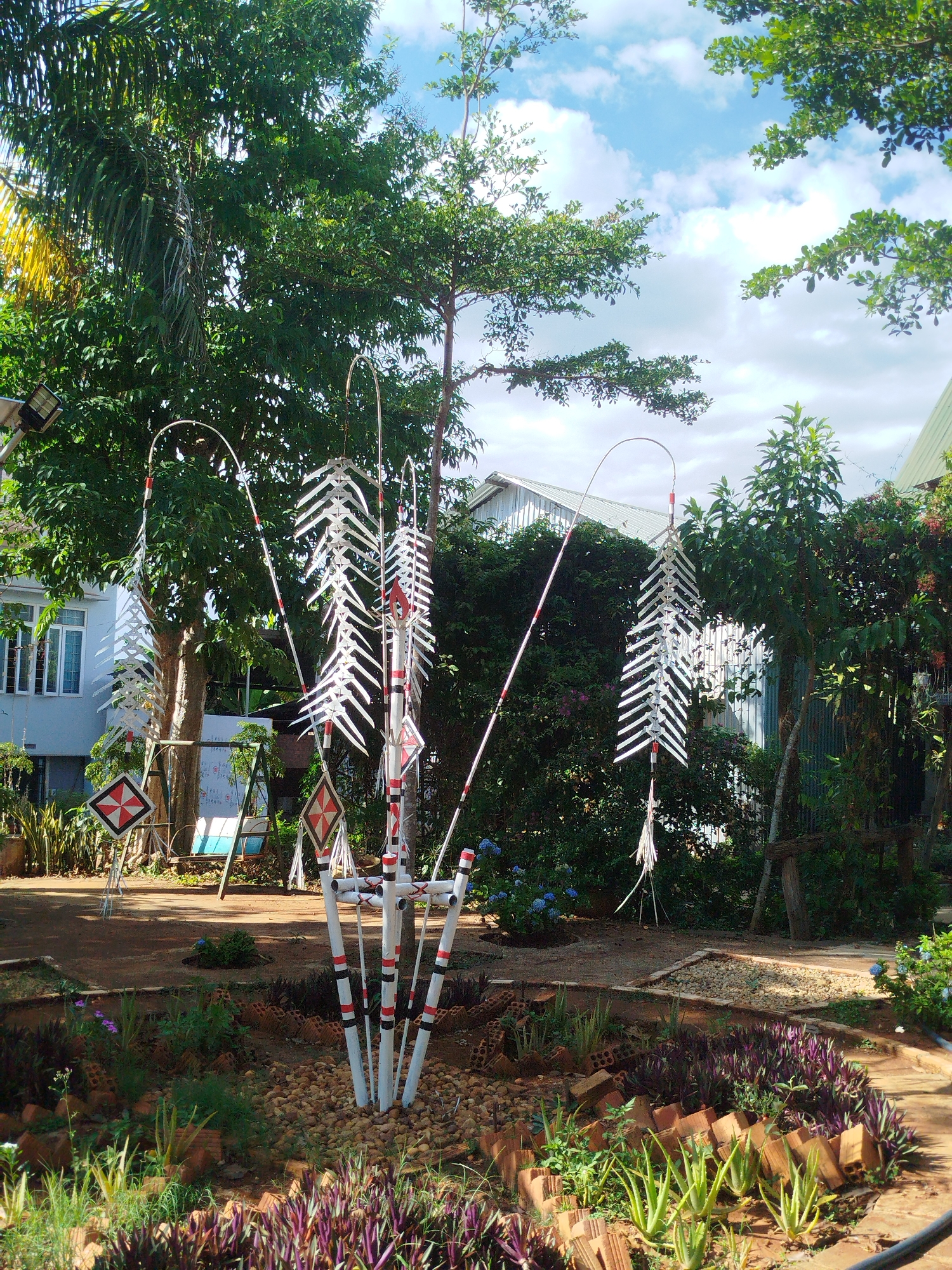 The image depicts a garden scene in An Phu Ward, Vietnam. In the center is a decorative structure consisting of multiple white poles banded with red and black stripes. Above the poles, curved metal rods extend outwards, each supporting a hanging ornament of stacked white panels. The poles are rooted in a small, raised bed surrounded by various plants including aloe vera and purple groundcover. The bed is bordered by brick and small stones.
In the background are lush green trees and vegetation. A white building with multiple windows is partially visible on the left, and a glimpse of a silver corrugated roof can be seen further back. An outdoor swing set and several diamond-shaped signs on poles are placed on the left. The overall scene is bathed in daylight, with a partly cloudy sky visible through the trees.