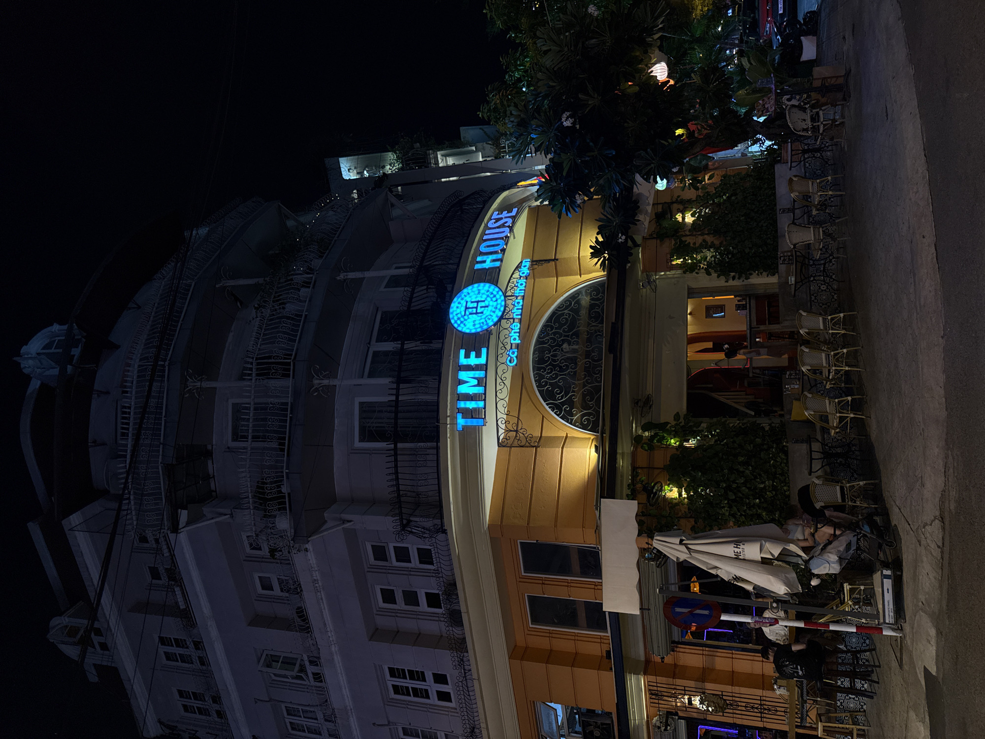 A street-level view at night of a building with a bright orange facade and a light gray upper section, likely a restaurant or cafe, in Nha Trang, Vietnam. The building is illuminated by neon signage that reads "TIME HOUSE" in blue letters. Below the main signage, smaller text reads "Ca phe - nha hang - thoi gian" (Coffee - restaurant - time) in a lighter color. An arched architectural feature with decorative metalwork is above the entrance. The street in front of the establishment is paved, with several tables and chairs set up on the sidewalk. A large white umbrella is positioned near the entrance, obscuring some of the view of the seating area. People are visible seated at tables inside and outside. There are trees and foliage surrounding the building, with decorative lanterns hanging from branches. The street is dark, with the primary illumination coming from the building's signage and interior lights. A red car and a motorcycle are parked on the right side of the image. The overall scene suggests a lively evening at a popular spot.