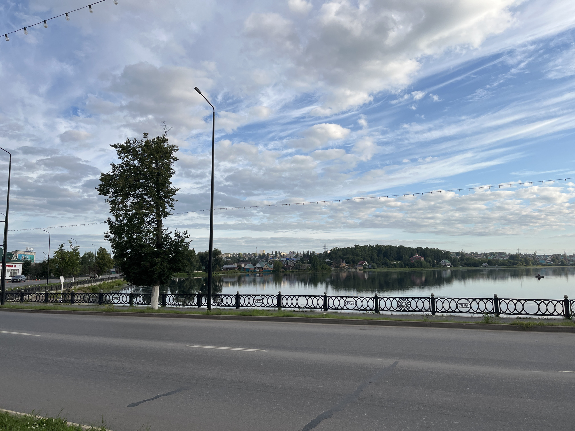 The image depicts a street scene alongside a body of water, likely a river or lake, in Blagoveshchensk, Russia. In the foreground, a two-lane asphalt road with white lane markings stretches across the frame. A dark, oil-like stain is visible on the right side of the road. Bordering the road and the water is a black metal fence with an ornate, circular pattern. Small patches of grass grow along the edge of the road and behind the fence.
Beyond the fence, a calm body of water reflects the sky and the opposite shore. Along the far bank of the water, a densely wooded area is dotted with houses, suggesting a residential area. The sky is filled with a mix of white and gray clouds, with patches of blue sky visible. Several streetlights with exposed bulbs are strung across the top of the frame, and taller, modern streetlights stand along the embankment. A large, leafy tree is prominent on the left side of the water, and other smaller trees are scattered along the embankment. In the distance, a few buildings and what appears to be a tower are visible on the horizon. There are no people or vehicles directly visible in the main portion of the image, though a car can be seen in the far distance on the left side of the embankment. A small boat is visible on the water in the right mid-ground.