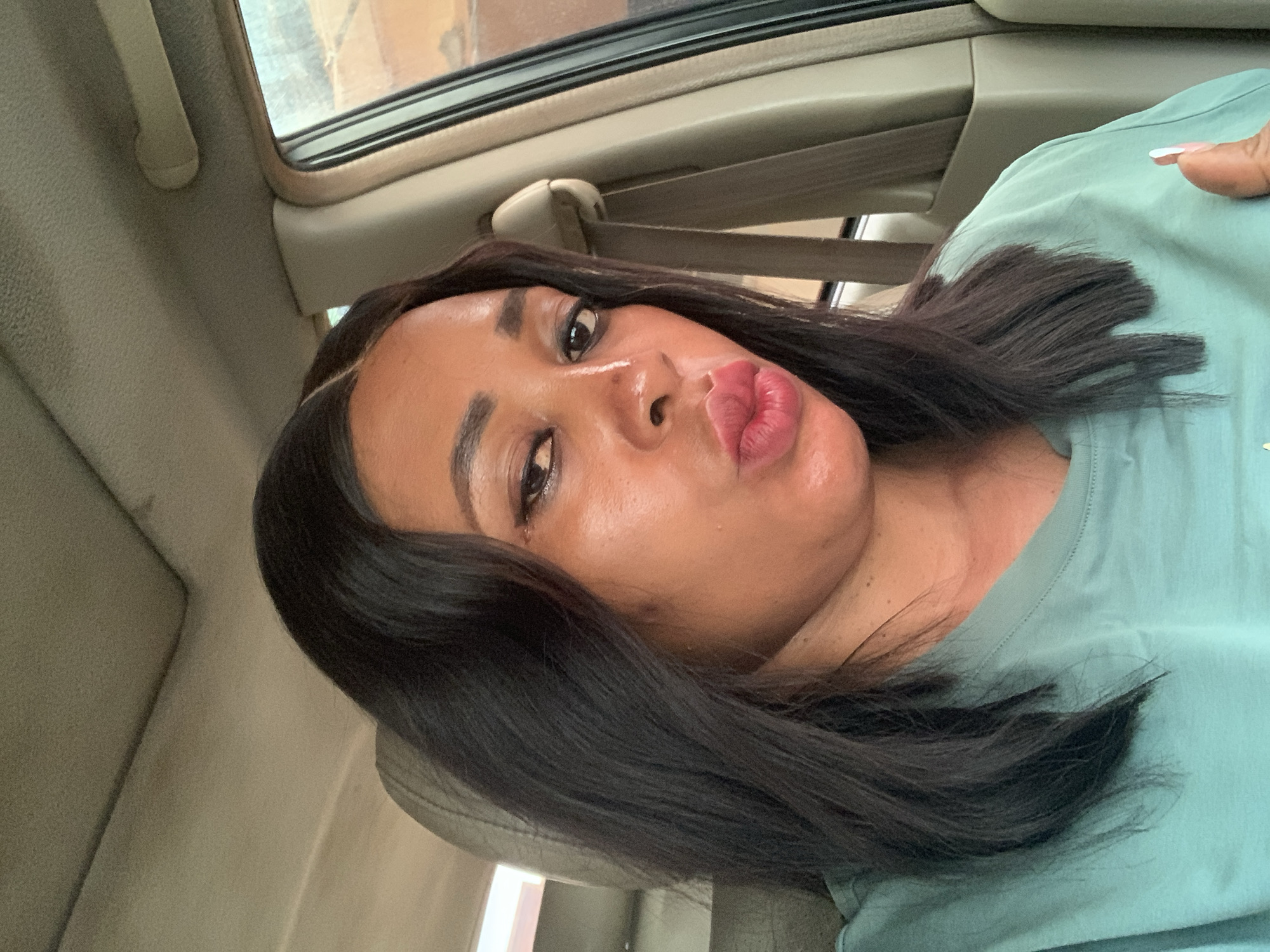 A woman with dark hair is posing for a selfie inside a car. The interior of the vehicle is visible, including the seat, seatbelt, and window. The environment is consistent with a car interior, no specific details identify the location in Sabon Tasha, Nigeria.