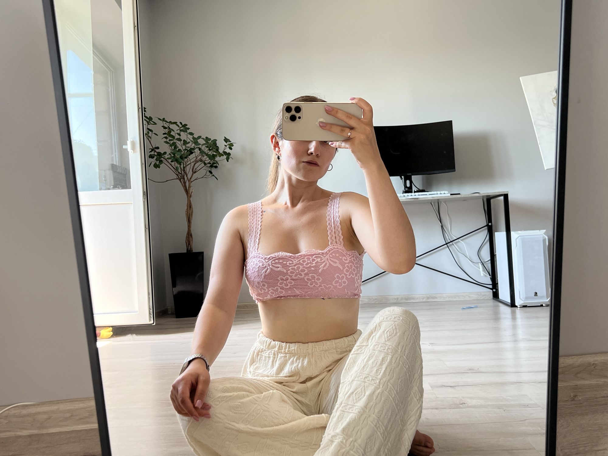 A young woman is sitting cross-legged on a wooden floor, facing a large mirror. She is taking a selfie with her phone, which is held up to obscure her eyes. She is wearing a pink lace bralette and loose, cream-colored pants with a textured pattern. A silver wristwatch is visible on her left wrist. In the reflection, behind her, is a desk with a black computer monitor and keyboard, a white computer tower, and some cables. To the left of the desk, a tall, dark pot with a small tree is visible. To the far left of the reflection, part of a large window or glass door can be seen. The room appears to be a bedroom or living area with light-colored walls and light wood flooring. This scene is consistent with a domestic setting within the city of Ternopil, Ukraine.