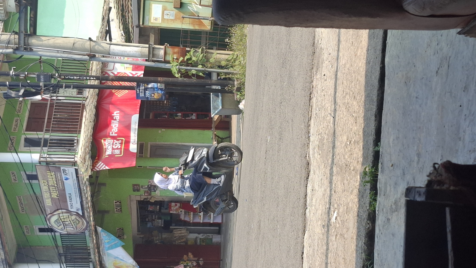 The image captures a street scene in Cilaku, Indonesia, with a young woman wearing a hijab and a patterned shirt riding a dark-colored scooter. She is positioned in the middle of a paved road, with her right hand on the scooter's handlebar. The scooter has a black body and silver accents.
In the background, a vibrant green building with multiple windows and balconies dominates the scene. A large red banner with white text that reads "TOKO SRC Fadilah" is prominently displayed on the building's facade. Above this, another banner in earthy tones features the word "JUARA" in a circular emblem. There are also various store entrances and displays visible along the building's ground floor.
A utility pole with wires runs vertically near the right side of the frame. To the right of the pole, a green electrical enclosure box is visible. The road surface is asphalt, and there is a concrete curb and sidewalk area. Small plants and weeds grow along the edge of the sidewalk. In the foreground, a portion of a dark, textured object, possibly furniture or a wall, partially obscures the bottom of the image. The lighting suggests it is daytime, with shadows cast by the structures and the scooter.