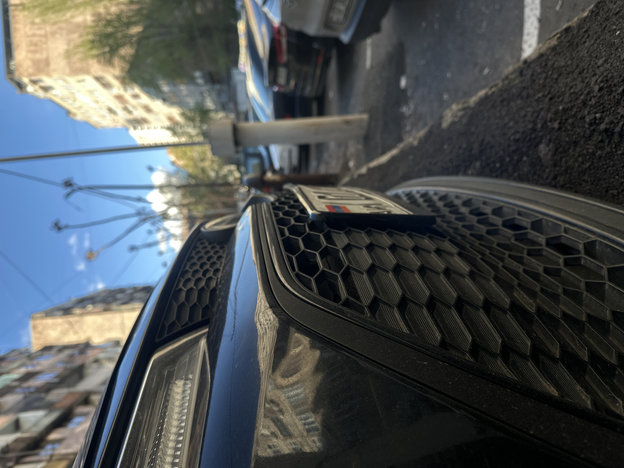 The image is a close-up shot taken at ground level, focusing on the front of a black car parked on a street. The car's front grill, featuring a honeycomb pattern, is prominently displayed, with a license plate affixed to it. Behind the car, another vehicle is partially visible. The surrounding environment includes a city street with asphalt pavement and a concrete curb. In the background, buildings of varying heights are seen, along with utility poles and wires against a blue sky with some clouds. The overall scene suggests an urban setting on a sunny day.
