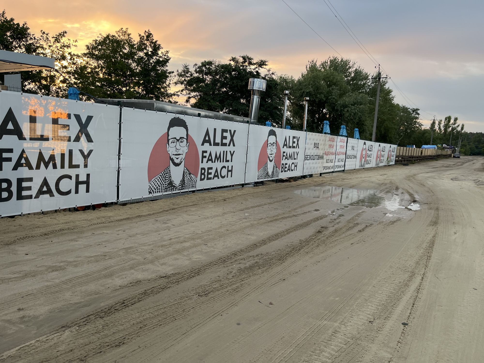 This media file depicts an outdoor scene at what appears to be a beach or recreational area. The dominant feature is a long, white banner stretching across the frame, featuring large black text that reads "ALEX FAMILY BEACH" multiple times. Interspersed with the text are stylized portraits of a man wearing glasses. The banner is supported by poles, and in some sections, Cyrillic text is visible, including words like "ВХІД" (entrance) and "БЕЗКОШТОВНИЙ" (free).
The ground in the foreground is sandy and shows tire tracks from vehicles, suggesting it might be a pathway or parking area leading to the beach. Several puddles of water are visible on the sand, reflecting the sky and surrounding structures. Behind the banner, trees and some industrial-looking structures, possibly part of a building or facility, are present.
The sky above is filled with dramatic clouds, illuminated by a warm, soft light, indicating either sunrise or sunset. The colors of the sky range from pale yellow and orange to dusky pink and grey. The overall atmosphere is serene, possibly hinting at the end of a day at the beach or an approaching change in weather. The scene is set in Bezliudivka, Ukraine, as indicated by the location context.