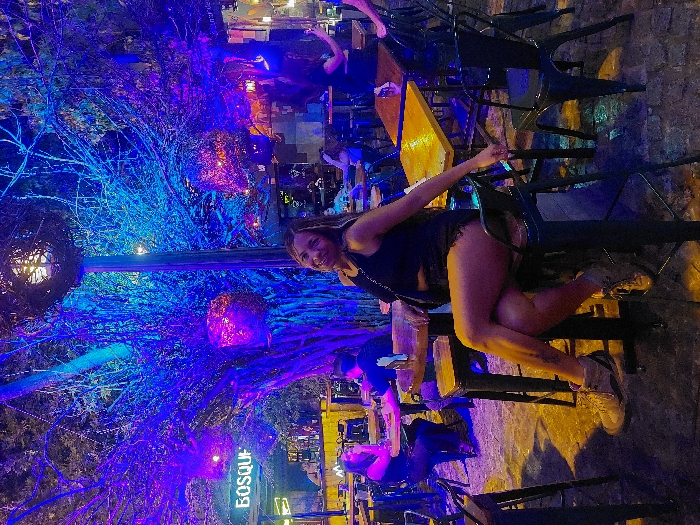 A woman sits on a bar stool at an outdoor venue called BOSQUE, identified by signage. She wears a black top and shorts with tan boots. She smiles and leans back with one arm on the chair next to her. The venue is lit with blue and purple lights accentuating tree branches and decorative spherical fixtures. Other people are visible in the background at tables.