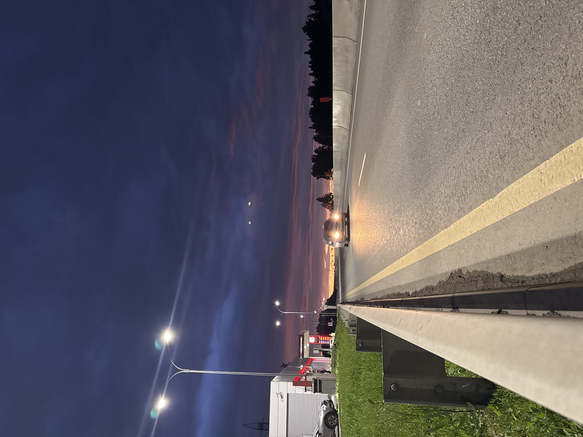 This vertical, low-angle, eye-level shot depicts a roadside scene at dusk, likely in or near the city of Можга, Russia. The foreground features a concrete barrier and a grassy verge with a metal guardrail. The asphalt road stretches into the distance, marked by a yellow line and a white line. A single car with its headlights on is driving on the road. In the mid-ground, a gas station with illuminated pumps and a building is visible on the left, with a parked car beside it. The sky is a deep twilight blue, transitioning to shades of pink and orange near the horizon, with some scattered clouds. Two streetlights with visible bulbs illuminate the scene. The background consists of a dark line of trees.