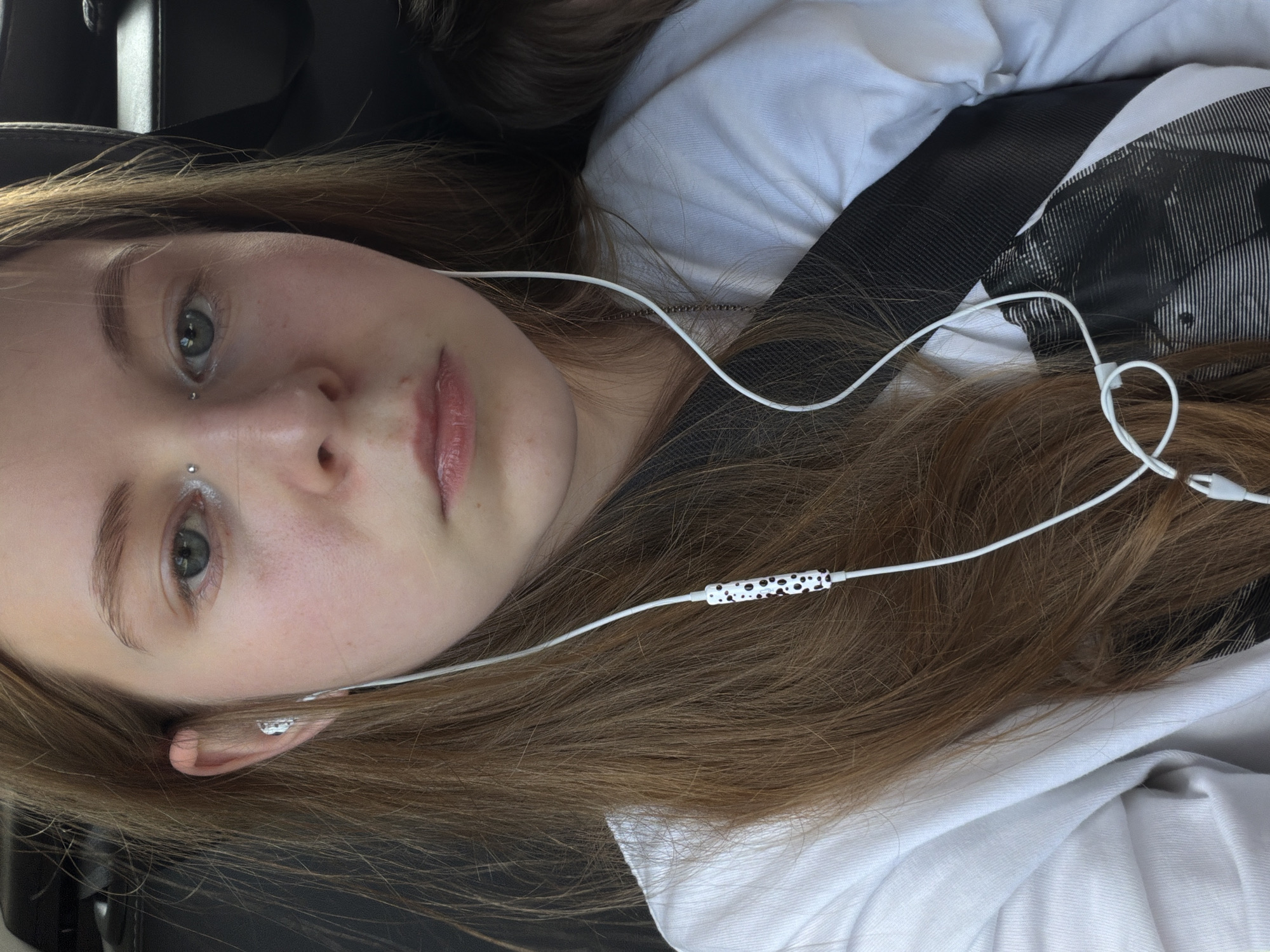 A close-up shot shows a young Caucasian female with long, light brown hair and blue eyes. She is wearing a white long-sleeved shirt and has earphones in her ears, with the white cord featuring a black and white spotted pattern. She also has a piercing on her bridge. The background appears to be the interior of a vehicle. The image was taken in Novoivanovskoe, Russia.