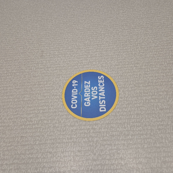 The image displays a circular blue and yellow sticker affixed to a light beige wall with a subtle, textured, repetitive pattern. The sticker, centered in the frame, features a yellow outer ring. Inside, a blue background contains white text. The top line reads "COVID-19". Below this, a thin yellow horizontal line is interrupted by a small upward-pointing yellow triangle, symbolizing distance. Beneath the line, the text "GARDEZ VOS DISTANCES" is displayed in white, which translates to "KEEP YOUR DISTANCES." The wall's texture appears to be a stylized, light-on-light pattern resembling intricate script or abstract shapes. No people, actions, or interactions are visible. The scene is an indoor environment in Strasbourg, France; however, a specific location or address within the city cannot be identified from the visual information provided.