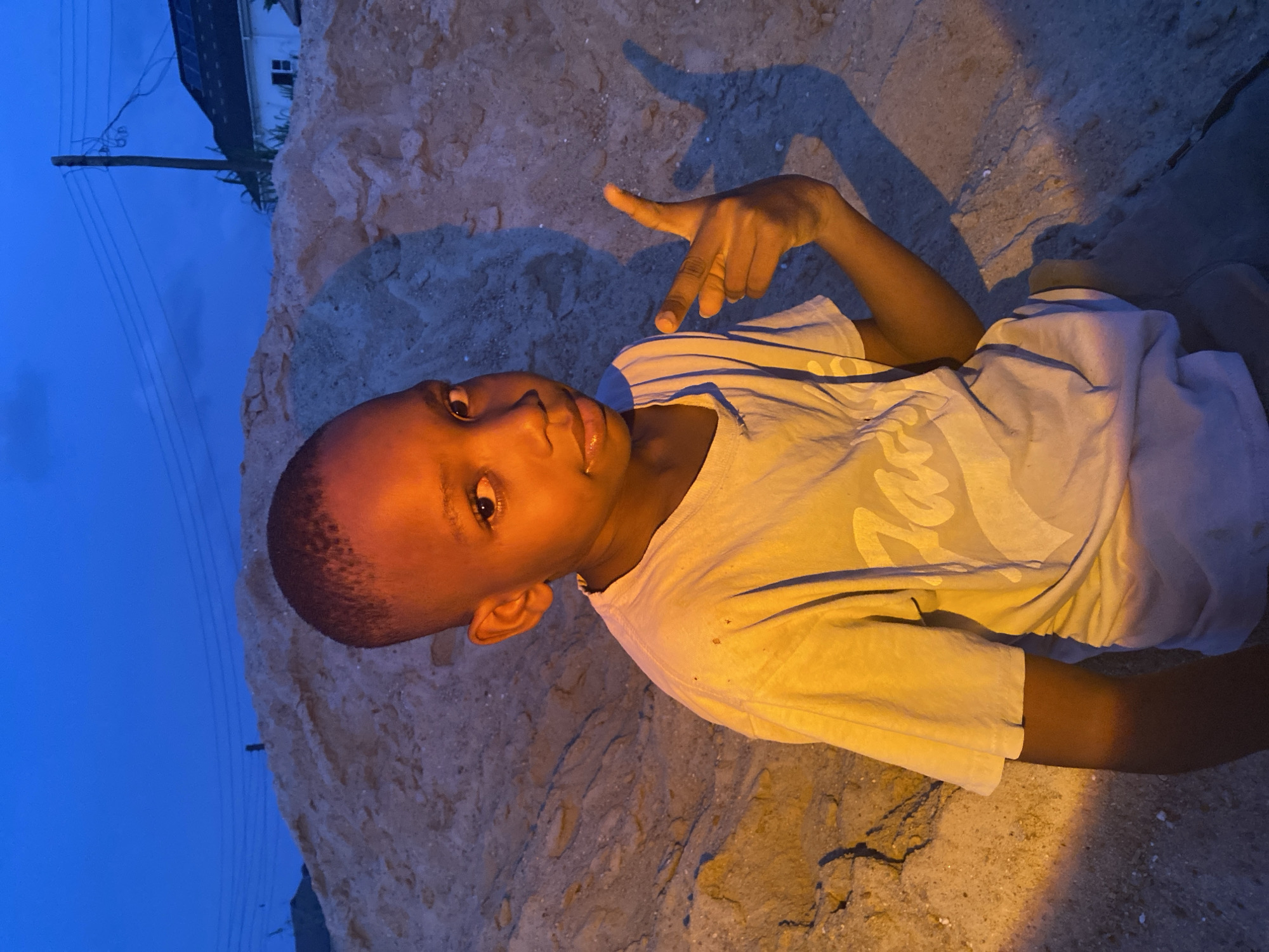 The image depicts a young male, approximately 7-10 years old, with dark skin and a short haircut, situated in Iranla, Nigeria. The scene is illuminated by a prominent warm-toned artificial light source from the lower left, indicating evening or nighttime conditions, contrasting with the deep blue sky. The individual is positioned centrally, facing the camera with direct eye contact, and appears to be sitting or kneeling on sandy ground. He is wearing a light-colored, short-sleeved t-shirt with faint graphic text and dark lower garments. His left hand is raised to chest level, with the index finger and thumb extended in a pointing gesture, casting a distinct blue shadow on the ground to his right. Behind the individual is a large mound of light-brown sand or dirt. In the background, a utility pole with multiple overhead wires is visible, along with a white residential building featuring solar panels on its dark-colored roof.