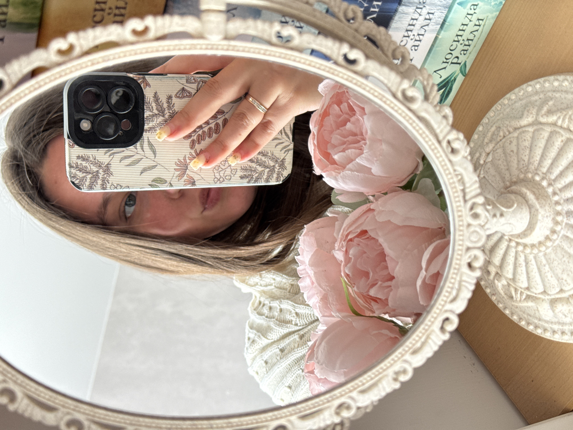 An indoor, close-up shot captures a person taking a mirror selfie. The central subject is the reflection of a person holding a smartphone, visible in an ornate, oval-shaped mirror with a cream-colored, carved frame. The person's face is partially visible, showing their eyes, nose, and mouth, framed by light brown or blonde hair. They appear to be looking directly into the phone's camera.
The smartphone is held by the person's right hand, revealing nails with a yellowish, possibly ombré or patterned, manicure. A silver or white gold ring with small stones is worn on the ring finger. The phone has a distinctive triple-lens camera array, suggesting a modern smartphone model, and is enclosed in a light-colored case adorned with a delicate botanical pattern of leaves and branches in muted earth tones.
To the right of the mirror, a cluster of soft pink, artificial flowers, resembling peonies or roses, adds a gentle touch to the scene, with some petals also reflected. Further to the right, a portion of another cream-colored, intricately carved decorative object, possibly a stand or base with a radial pattern, is visible. In the upper left background, as seen in the reflection, the spines of two books are stacked. The most prominent book spine clearly displays text in Cyrillic: "ЛЮСИНДА ДА РАЙЛИ" (Lucinda Riley). Partial Cyrillic text, "СИНДА ДА АЙЛИ," is visible on another book. The person is wearing a cream-colored, textured knitted garment, possibly a sweater. The lighting is soft and natural, suggesting daytime. The overall ambiance is cozy and personal.