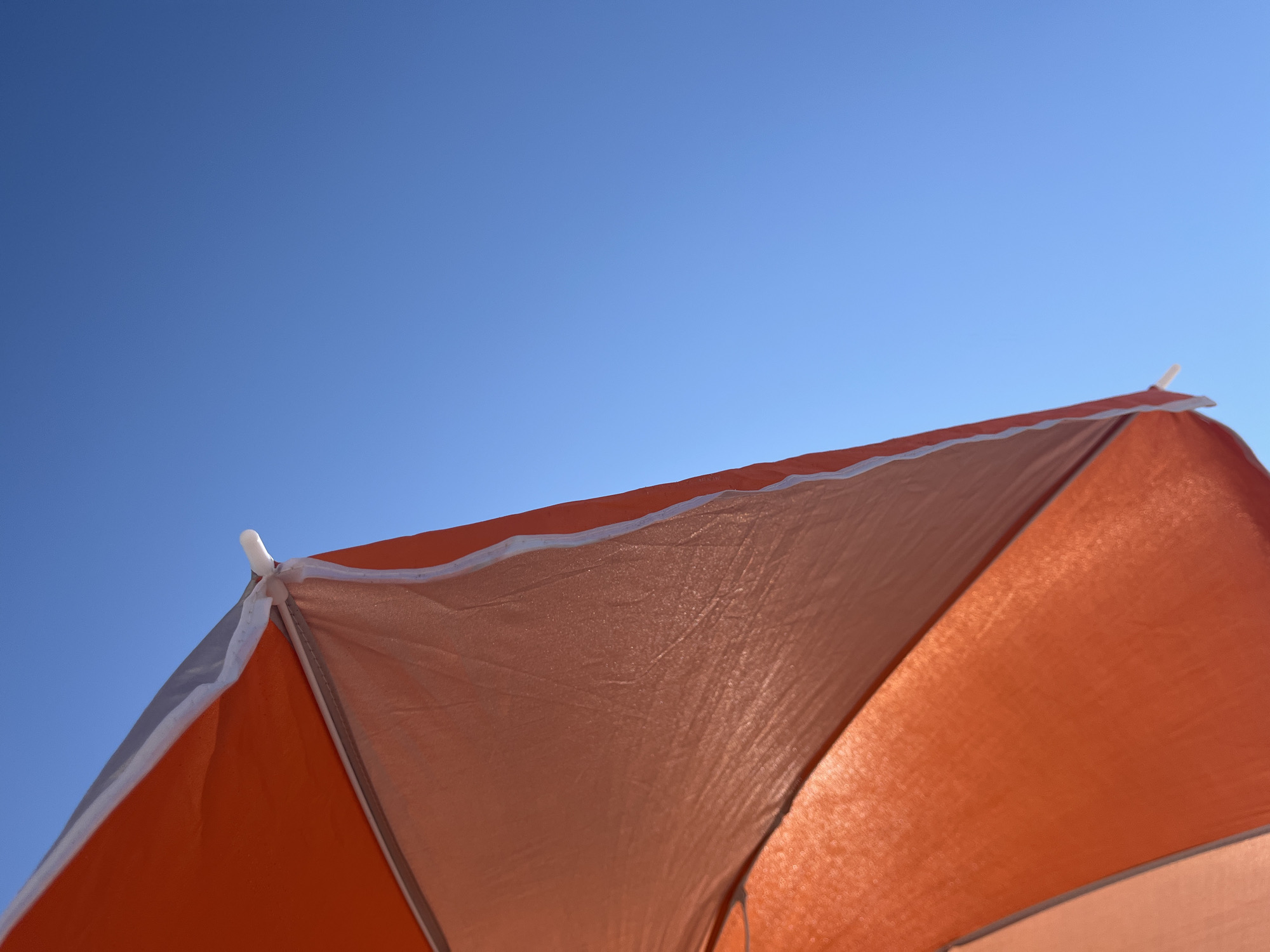 An orange and white beach umbrella is angled upwards against a clear blue sky. The umbrella's fabric is taut and slightly translucent, allowing sunlight to filter through. The top edge of the umbrella is visible, with a white trim. The bottom portion of the umbrella is a brighter orange, suggesting it is closer to the light source. The image is taken from a low angle, looking up at the sky and the umbrella. The location is Palavas-les-Flots, France, a coastal town known for its beaches.
