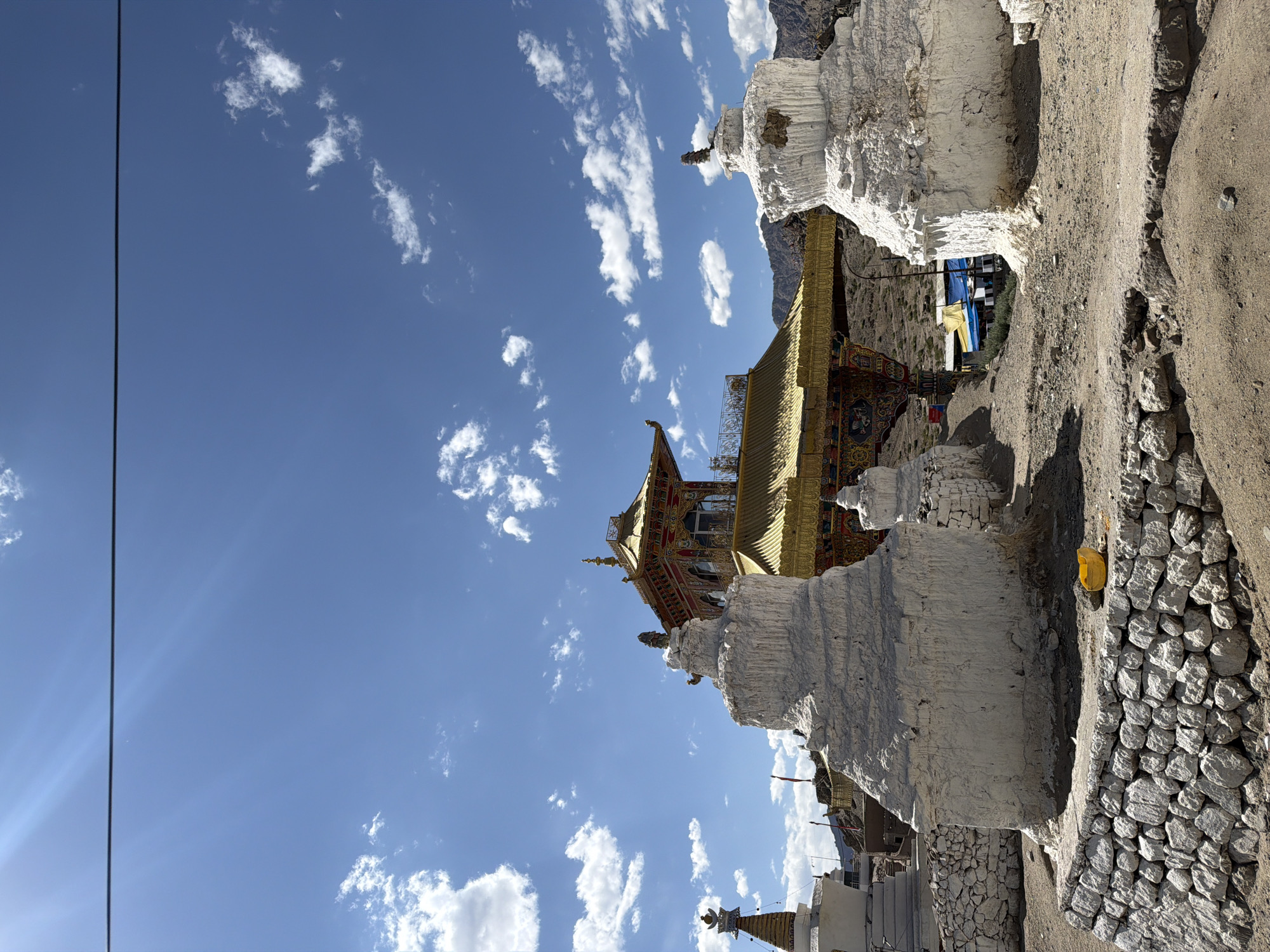 The image depicts a Buddhist monastery located in Leh, India, under a clear blue sky with scattered white clouds. In the foreground, two prominent white stupas, with ornate finials, stand on a rocky foundation. A low stone wall borders the area in front of the stupas. Behind the stupas, a vibrantly decorated golden gateway with intricate carvings and Buddhist motifs leads to a larger, golden-roofed structure. The background shows barren, mountainous terrain. A single black wire stretches horizontally across the upper portion of the sky.