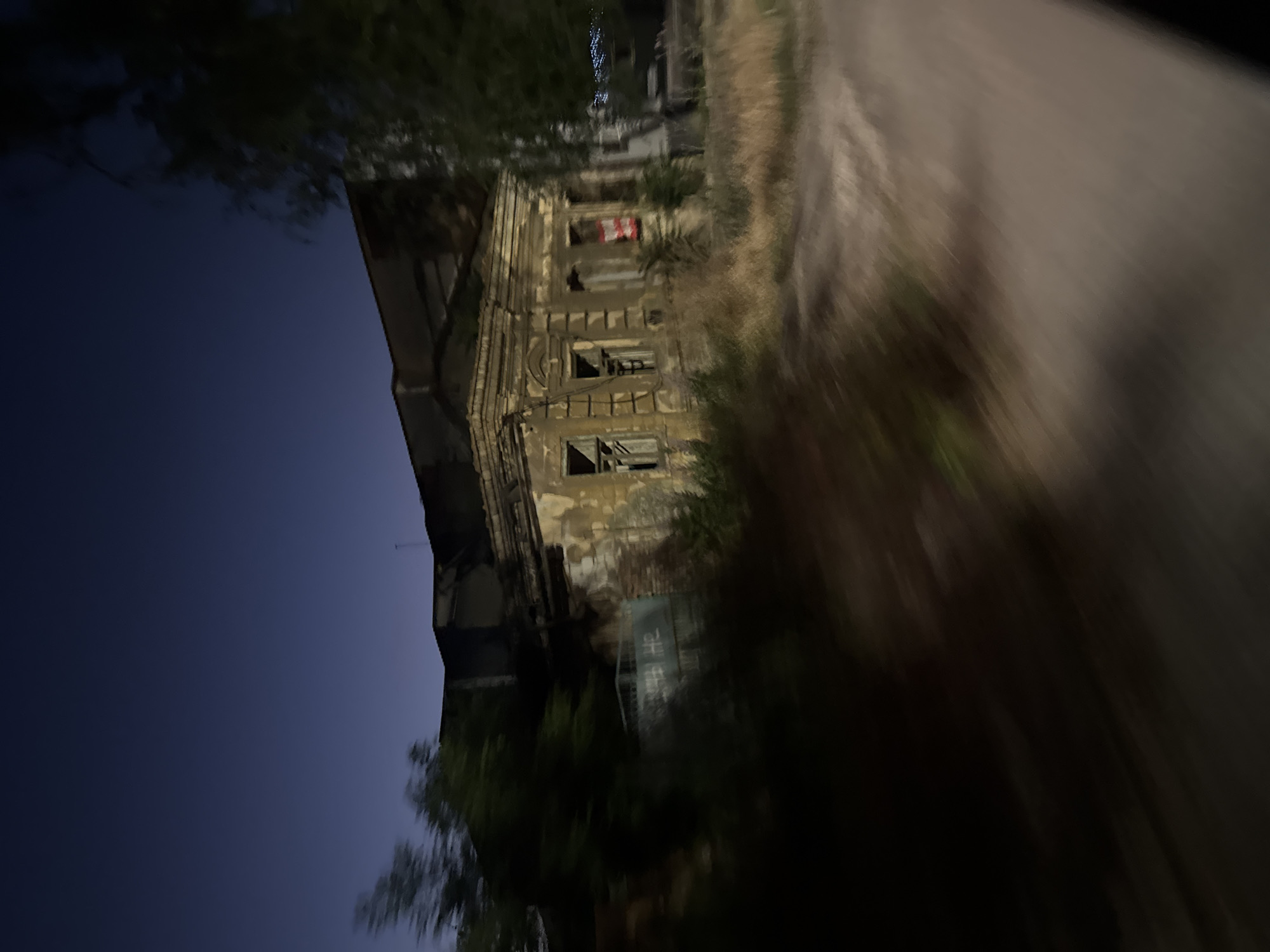 This image, captured at dusk in the city of Mariupol, Ukraine, depicts a partially ruined building with peeling yellow paint and visible brickwork. The structure features ornate architectural details, including pilasters and decorative cornices, suggesting it may have been a house of some significance. Several windows are broken or boarded up, and the surrounding area is overgrown with dry grass and bushes. A rusty metal gate with visible graffiti is present to the left of the building. The sky is a deep blue, indicating twilight. The image appears to have been taken from a moving vehicle, resulting in motion blur, particularly in the foreground vegetation and the road. No people are visible in the scene.