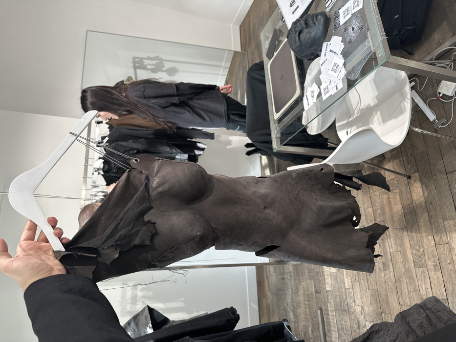 An interior scene depicts a contemporary fashion showroom in Paris, France. In the foreground, a person's light-skinned hand, clad in a black long-sleeved garment, holds a white wooden clothes hanger. Suspended from the hanger is a dark, distressed, leather-like garment sculpted to resemble a female torso, featuring distinct breasts and an abdominal section. This garment exhibits ragged, torn edges along its bottom and sides, with visible metal eyelets and thin cords near its top.
In the mid-ground, a person with long dark hair, wearing a black long-sleeved shirt and dark trousers, stands facing right, partially obscured by the foreground garment. A segment of a second individual's bald head is visible behind the held garment and to the left of the first person. Behind them, a metal clothing rack displays several dark-colored garments. A large, rectangular mirror leans against a white wall on the right side of the frame.
A clear glass-top table with a silver metal frame occupies the right mid-ground. Upon it are a silver laptop, a dark mobile phone, a black anatomical face mask, and several white rectangular cards, some featuring QR codes and the text "LIZA BEAMS." Two white, modern chairs with slender metal legs are positioned around the table. The floor is composed of light brown wooden planks arranged in a herringbone pattern, and the walls are painted white.