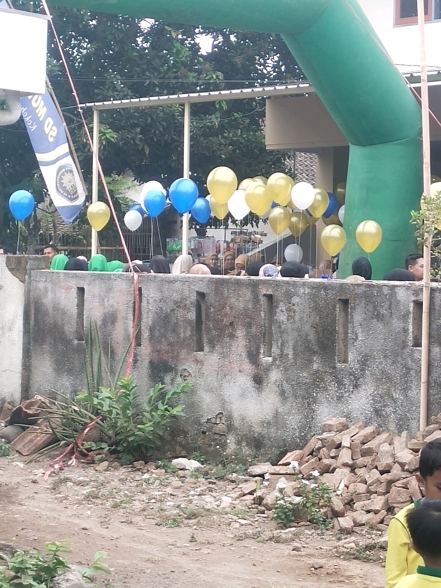 This image depicts an outdoor scene, likely in Ambulu, Indonesia, featuring a large green inflatable archway. Below the archway, a group of people are gathered, many wearing hijabs, standing behind a low concrete wall. The wall is textured and shows signs of wear. To the left of the arch, a blue and white flag is visible, partially unfurled. Numerous balloons, primarily blue and yellow, are suspended around the archway and above the gathered individuals, suggesting a festive occasion or event. In the foreground, the ground is unpaved and appears somewhat rough, with scattered debris, including bricks and small plants. The overall setting suggests a public gathering space, possibly near a school or community center, given the flag's inscription.