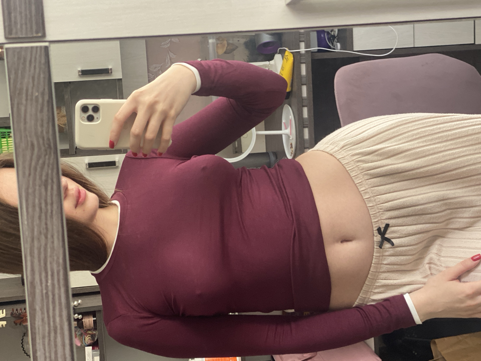 A full-torso mirror selfie shows a woman indoors, holding a white-cased smartphone with her right hand. She is wearing a form-fitting, long-sleeved burgundy top with white trim on the collar and cuffs, and light beige, pleated bottoms featuring a small black bow detail near the navel, which is visible. Her long brown hair is partially visible on the left side, and her lips are slightly parted. Both her visible hands have bright red nail polish.
The woman is positioned in front of a mirror with a decorative brown and light-colored frame. The mirror reflects parts of a room behind her, including light-colored cabinets with dark horizontal handles, a green storage basket, a small white stand-up fan, a yellow tube-like object, and a purple office chair. The lighting suggests an indoor setting, and no specific time of day is discernible. There is no visible text in the image. The overall impression is a casual, domestic scene.
