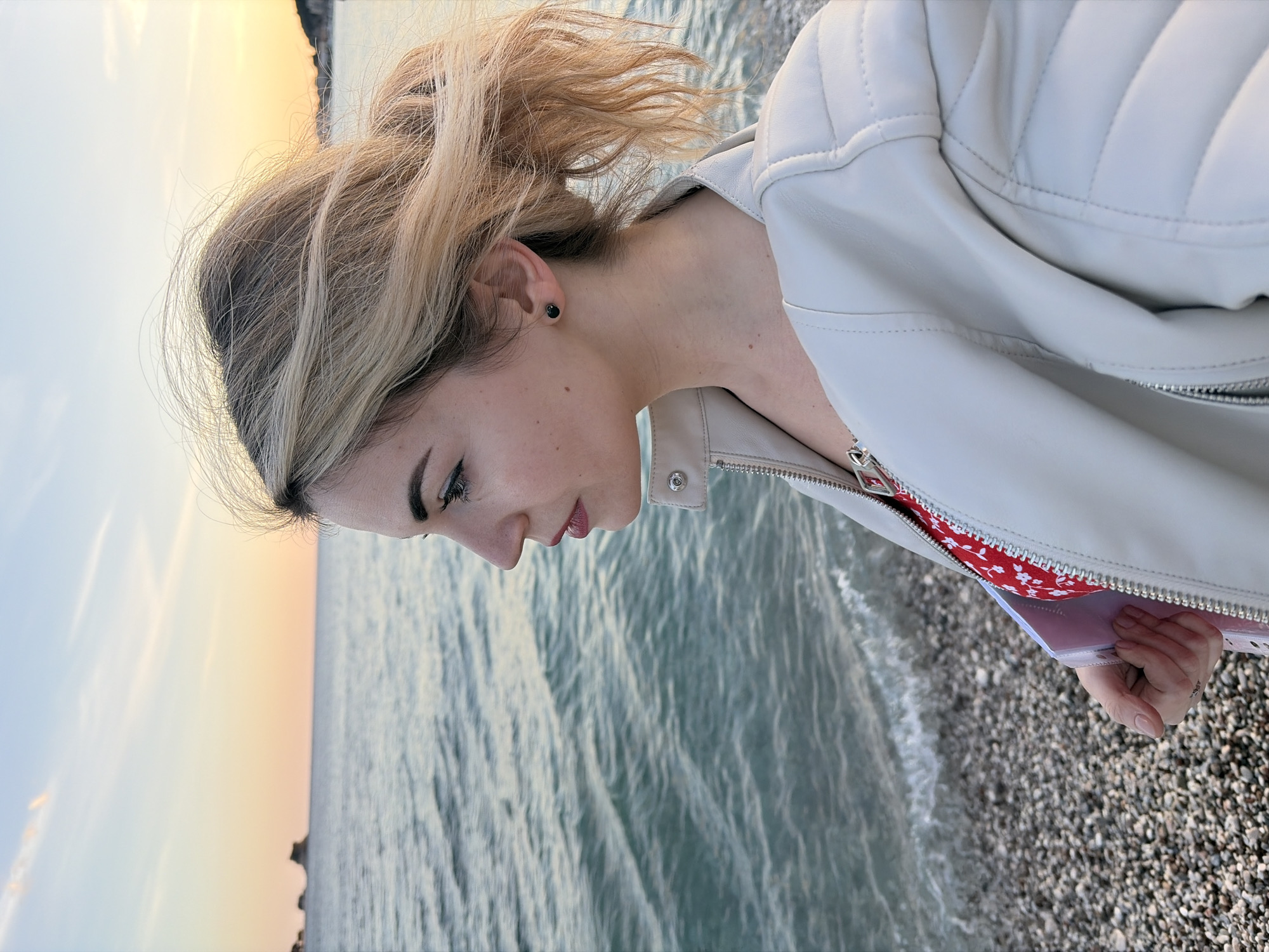 A female subject is depicted in profile, facing left, positioned on a pebble beach in Budva, Montenegro. The setting indicates either sunrise or sunset, with the sky displaying a gradient from light blue overhead to orange-pink at the horizon. The subject has blonde hair, partially windblown and gathered into a low ponytail, with a black stud earring in her visible left ear. Her facial features include defined eyebrows, eyeliner, and red-tinted lips. She wears an unzipped, light beige quilted leather jacket over a red and white patterned garment. Her left hand, partially obscured within the jacket, holds a white rectangular object. The foreground consists of a pebble beach composed of various grey and brown stones. The midground features the sea with small, turquoise-hued waves breaking on the shore. In the distant background, two rocky islets are visible in the sea to the left, and a faint coastal outline with structures appears on the right.