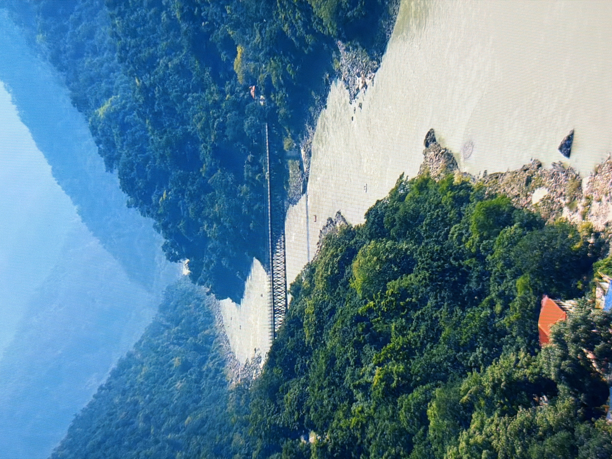 This aerial shot captures a wide river flowing through a lush, forested mountainous region. A striking suspension bridge, with a dark, latticed structure, spans the river, connecting the dense green slopes on either side. The river itself appears wide and a pale, milky color, with rocky banks visible in places. The landscape is dominated by verdant trees covering the steep hillsides. On the right bank, nestled amongst the trees, is a bright orange-roofed building, suggesting a small settlement or a structure within the natural landscape. The scene is bathed in what appears to be natural daylight, possibly mid-morning or late afternoon, given the soft illumination. The overall impression is one of a remote and natural setting, possibly a national park or a less developed area, characterized by its impressive river and the man-made structure of the bridge. There is no visible text in the image.