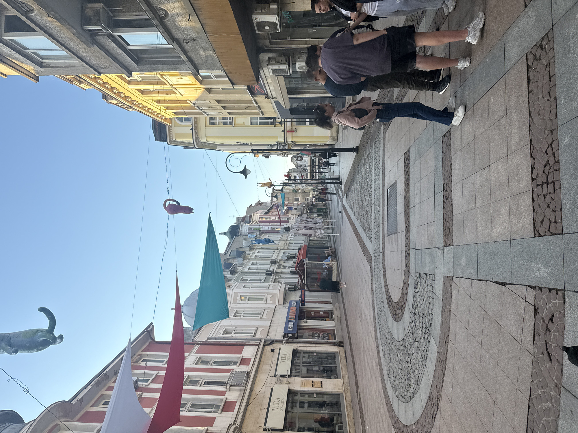The image shows a street view in Gabrovo, Bulgaria. The street is lined with buildings of varying heights and architectural styles, including shops and apartments. Overhead, there are colorful banners and decorations suspended on wires. People are walking along the paved sidewalks. The sky is clear and blue. The overall setting is a vibrant and urban pedestrian space.