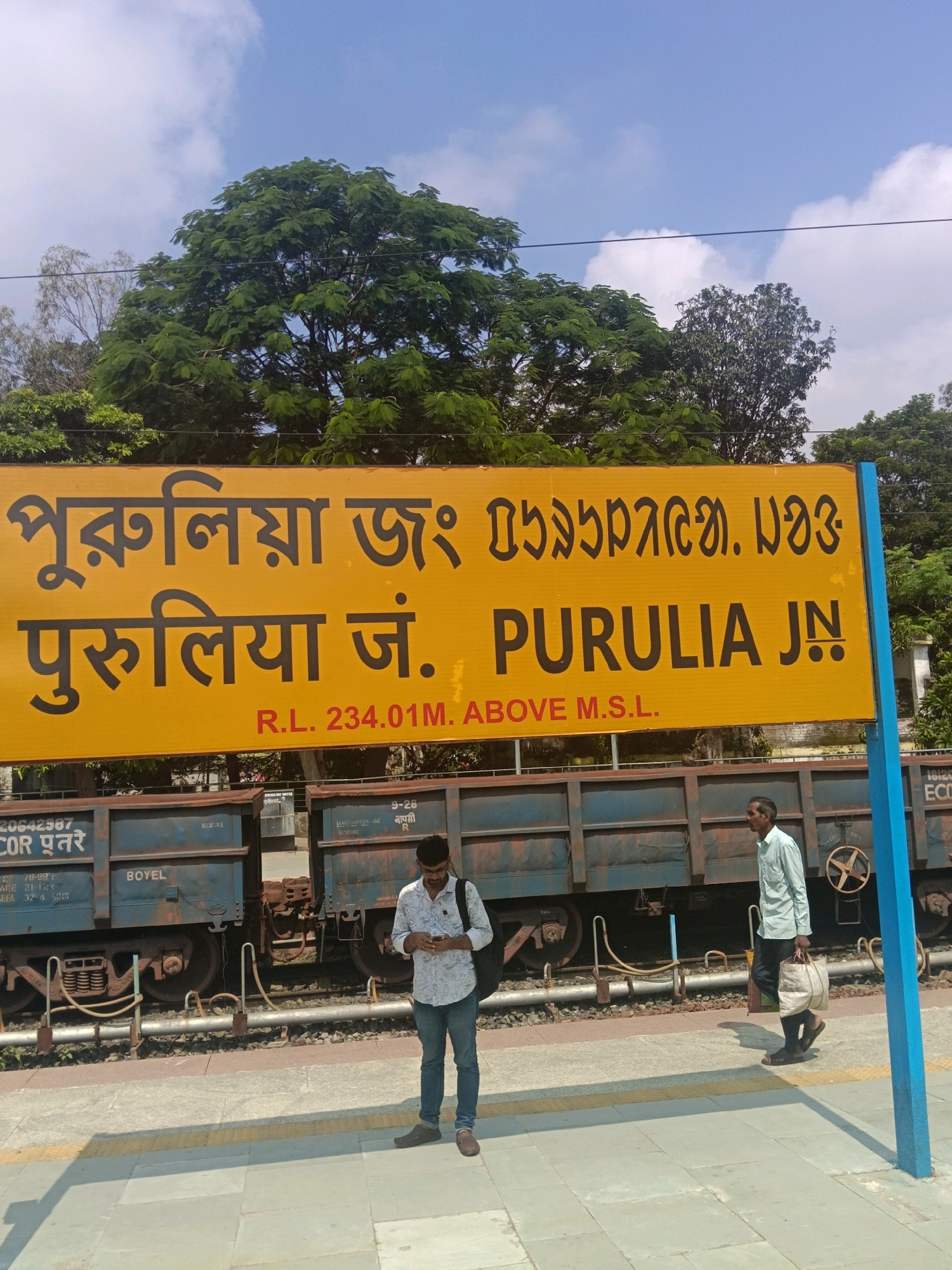 A yellow sign with black lettering in Bengali and English is mounted on a blue pole at a train station. The Bengali text reads "পুরুলিয়া জং" and the English text reads "PURULIA JN". Below this, in smaller red letters, it says "R.L. 234.01M. ABOVE M.S.L.". The sign is positioned in front of a train with blue carriages. Two men are visible in the frame. One man, wearing a white patterned shirt and blue jeans, stands on the platform, looking at his phone. Another man, wearing a light blue shirt and dark pants, walks past carrying a bag. The background shows trees and a blue sky with white clouds. The scene is a typical outdoor train station environment.