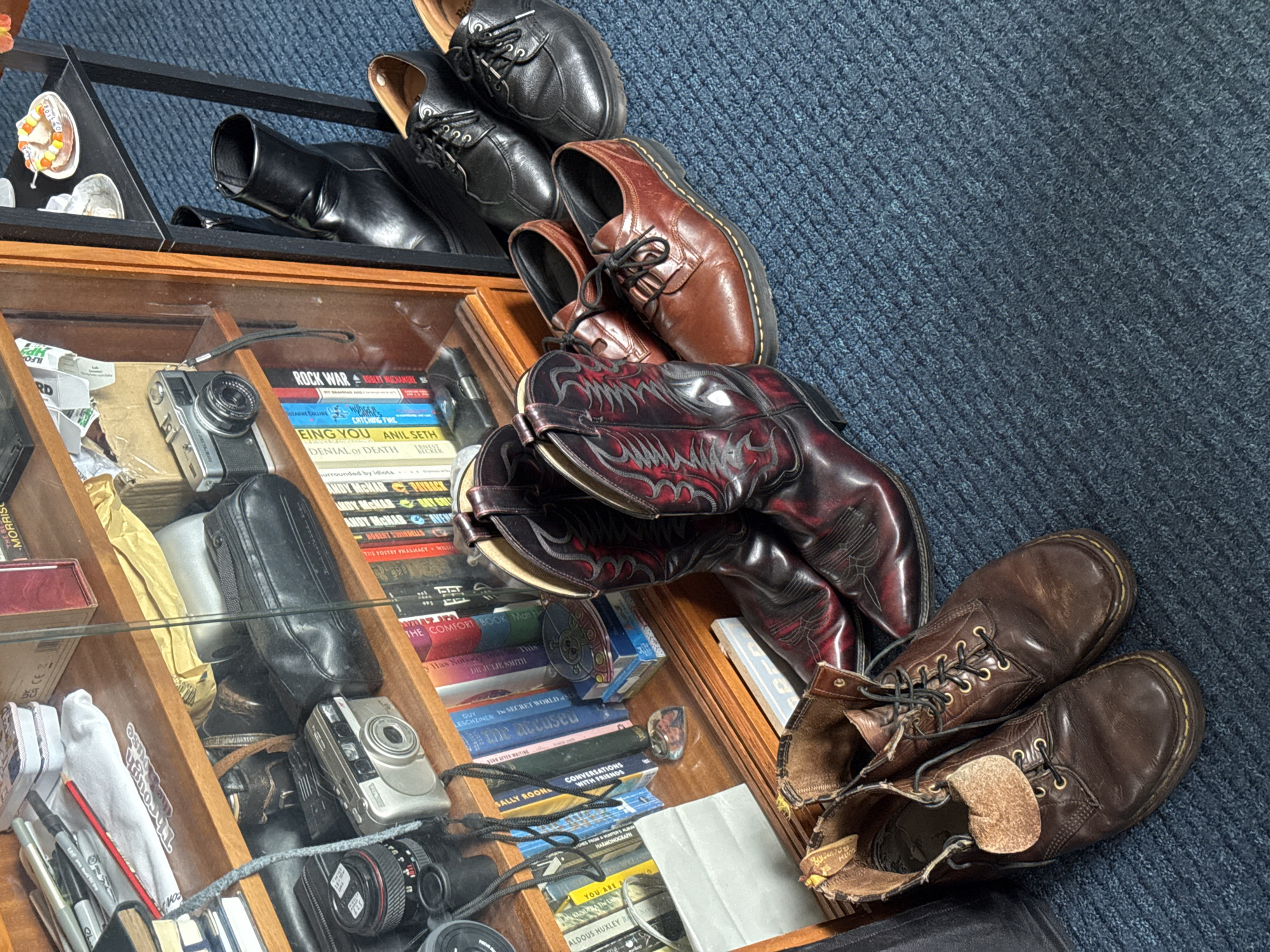 The scene is a glass-topped display case situated on a blue carpet. The case's contents include various pairs of shoes and boots: black dress shoes, brown leather shoes, red and black cowboy boots, and brown leather boots. Inside the case are books with titles like "Rock War," "Catching Fire," "Being You," "Denial of Death," and "Conversations with Friends," along with cameras, pens, pencils, and other miscellaneous items. A small tray in the upper left corner holds what appear to be candy corn.