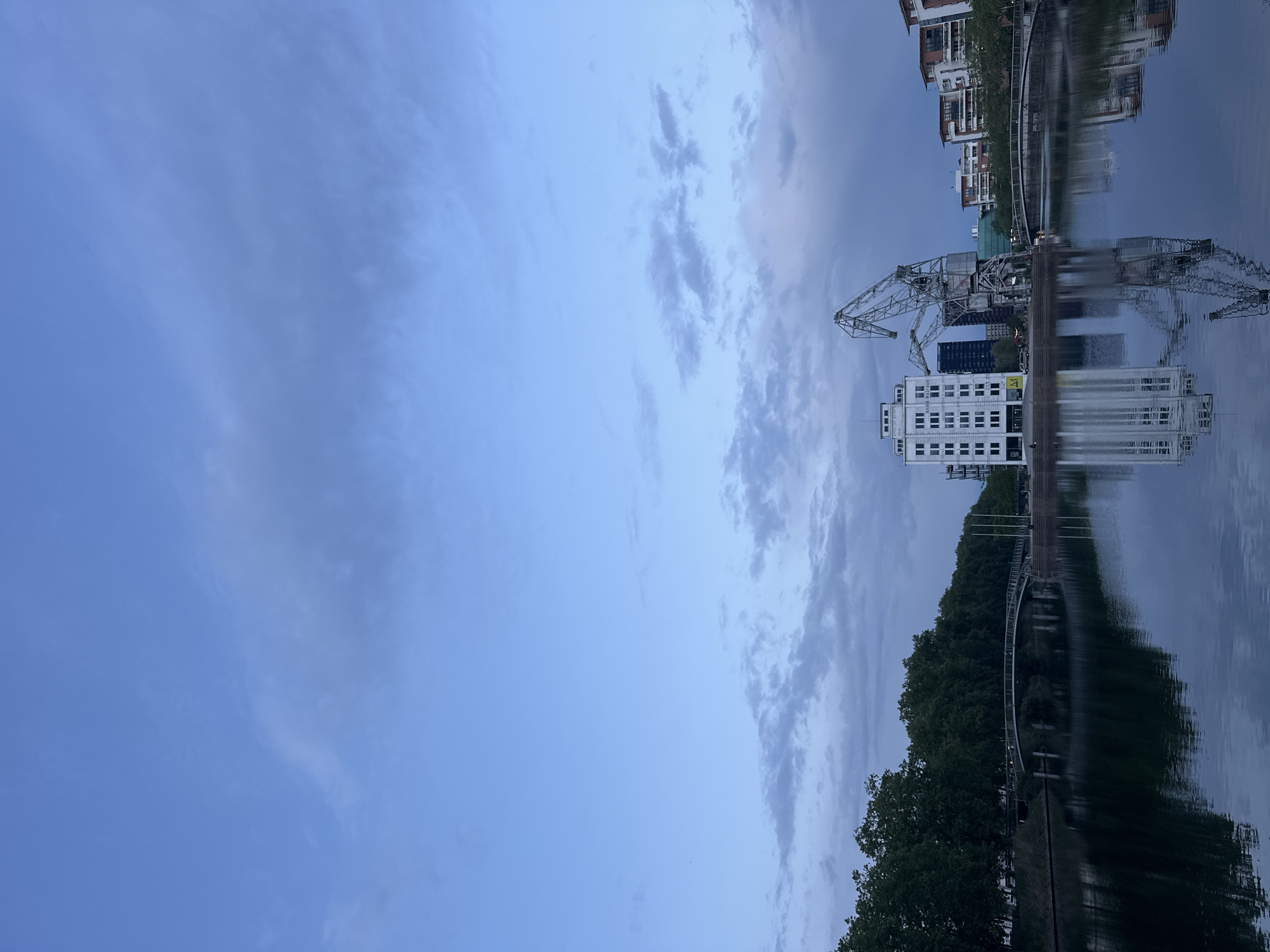 The image depicts a waterway, likely a canal or river, in Strasbourg, France, reflecting the sky and surrounding architecture. Dominating the skyline is a crane, possibly historic, alongside a modern white building with a grid of windows. Further along the water, there's a bridge or walkway lined with trees, and in the background, residential buildings are visible. The sky is overcast with patches of blue visible, suggesting an evening or early morning scene. The reflection in the water is clear, mirroring the structures above.