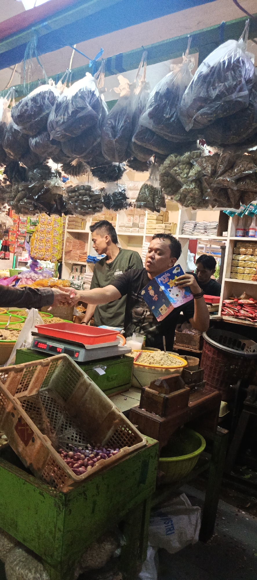 In Sukaresmi, Indonesia, a man in a dark t-shirt with a "Purple Cat" book is engaged in a transaction, possibly a sale, with another person whose hands are visible and shaking the vendor's hand. The vendor stands behind a counter in a market setting, with a digital scale displaying "00.00" and "44" prominently in the foreground. To the left, a green crate is filled with red onions, and another crate, partially obscured, also contains produce. Shelves in the background are stocked with various goods, including what appear to be bags of dried food items and packaged snacks. Bags of produce are hanging from the ceiling, suggesting a traditional market environment. Two other men are present, one in a green t-shirt looking towards the interaction, and another man partially visible in the background. The lighting is artificial, indicating an indoor or covered market.