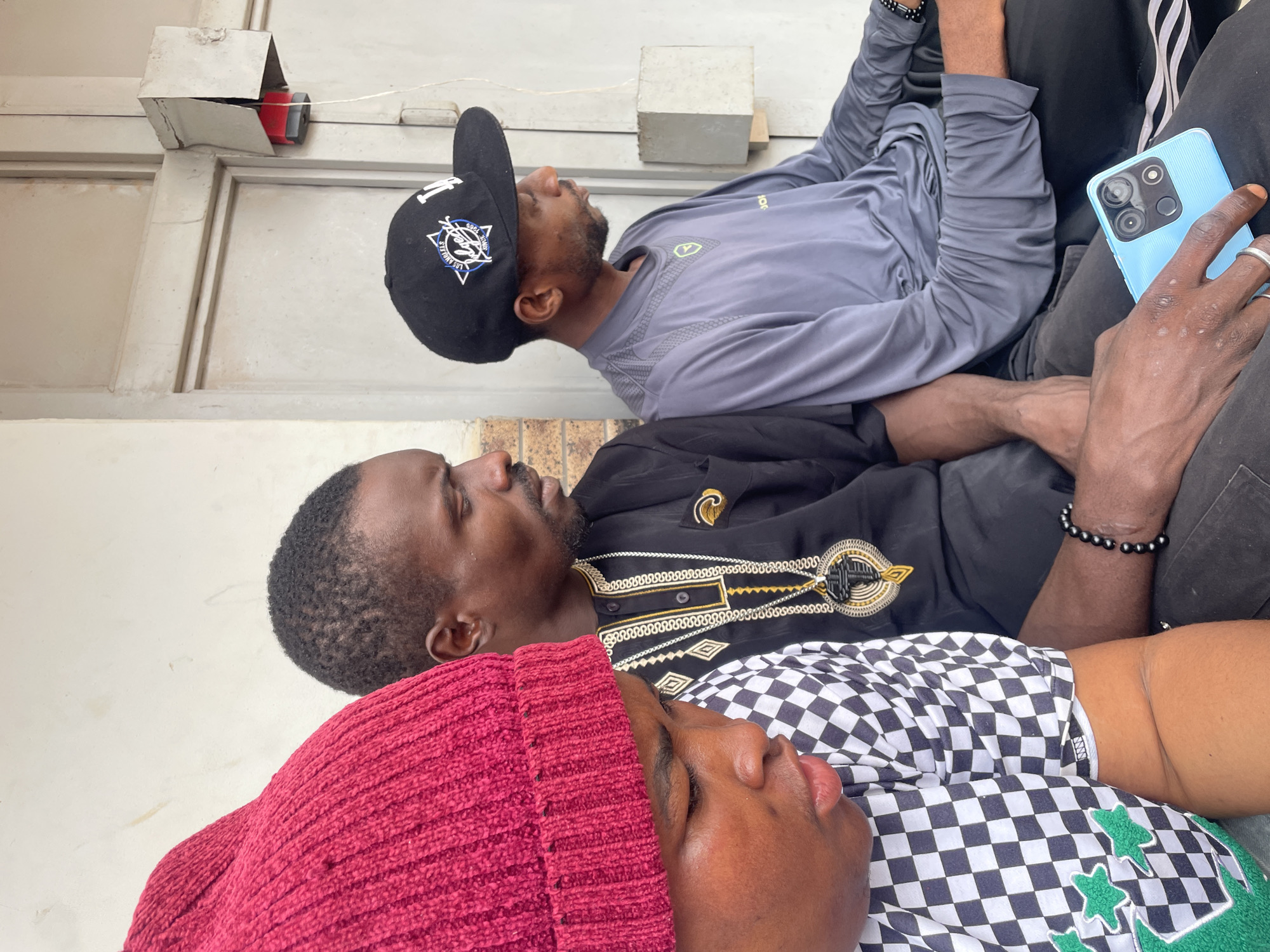 Three individuals are seated side-by-side in an outdoor setting, likely in Kurunduma, Nigeria. The person on the far left is wearing a red knitted hat and a black and white checkered shirt. To their right, a man is dressed in a black traditional garment adorned with gold trim and a prominent gold necklace featuring a cross pendant. He also wears a beaded black bracelet. The individual on the far right is wearing a grey long-sleeved shirt and a black baseball cap. A blue smartphone is held in the lap of the person on the right. The background consists of a light-colored wall and a metal doorway with a visible red alarm box mounted on it. The overall scene suggests a moment of pause or waiting.