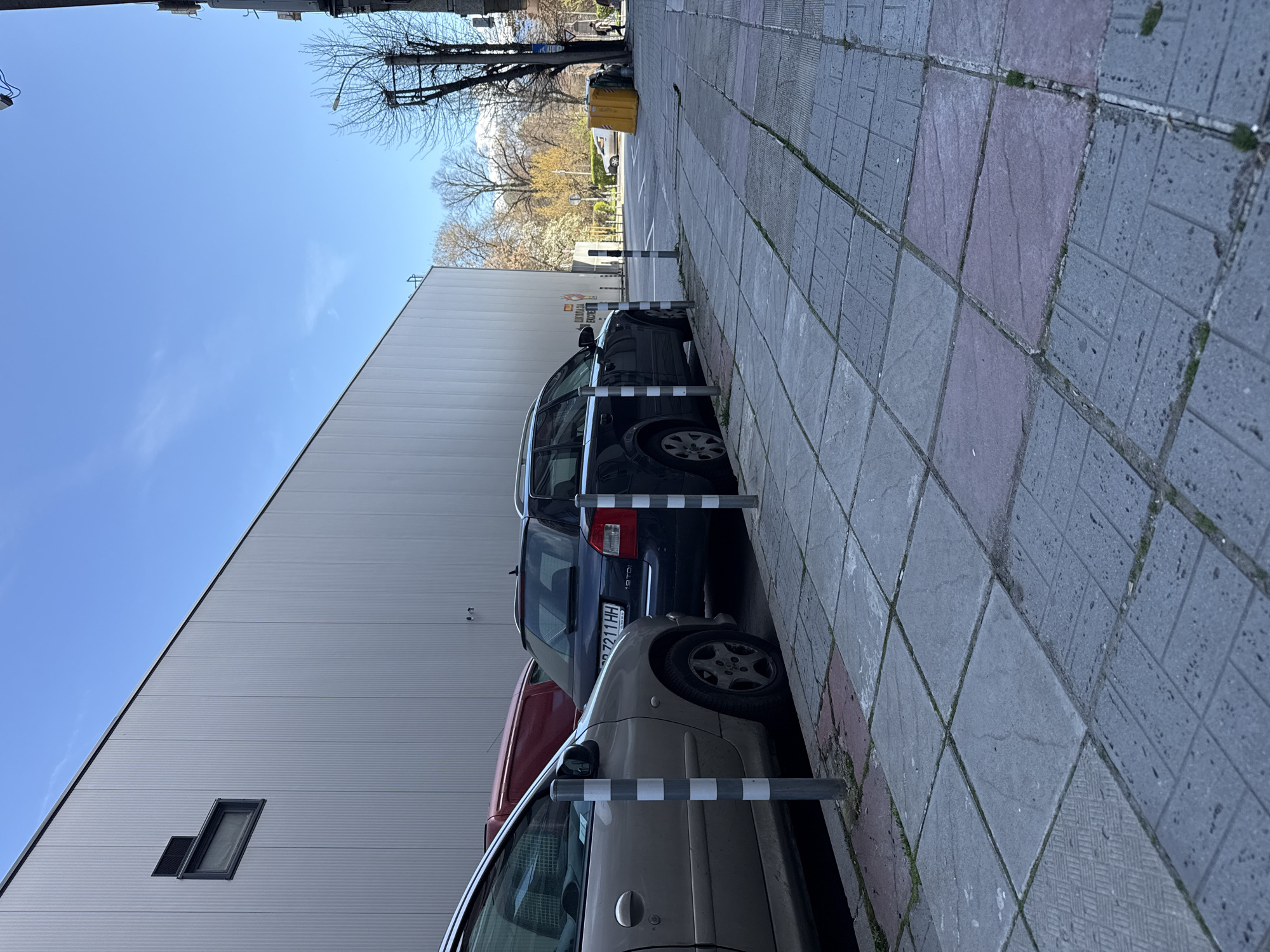 A street scene in Unknown, Unknown shows parked cars aligned between striped posts and the side of a large, gray building. The building has a few small, rectangular windows near its base. The sky is a clear, bright blue. A sidewalk with gray and pink pavers runs parallel to the cars and building. In the distance, trees with bare branches and a yellow dumpster are visible.