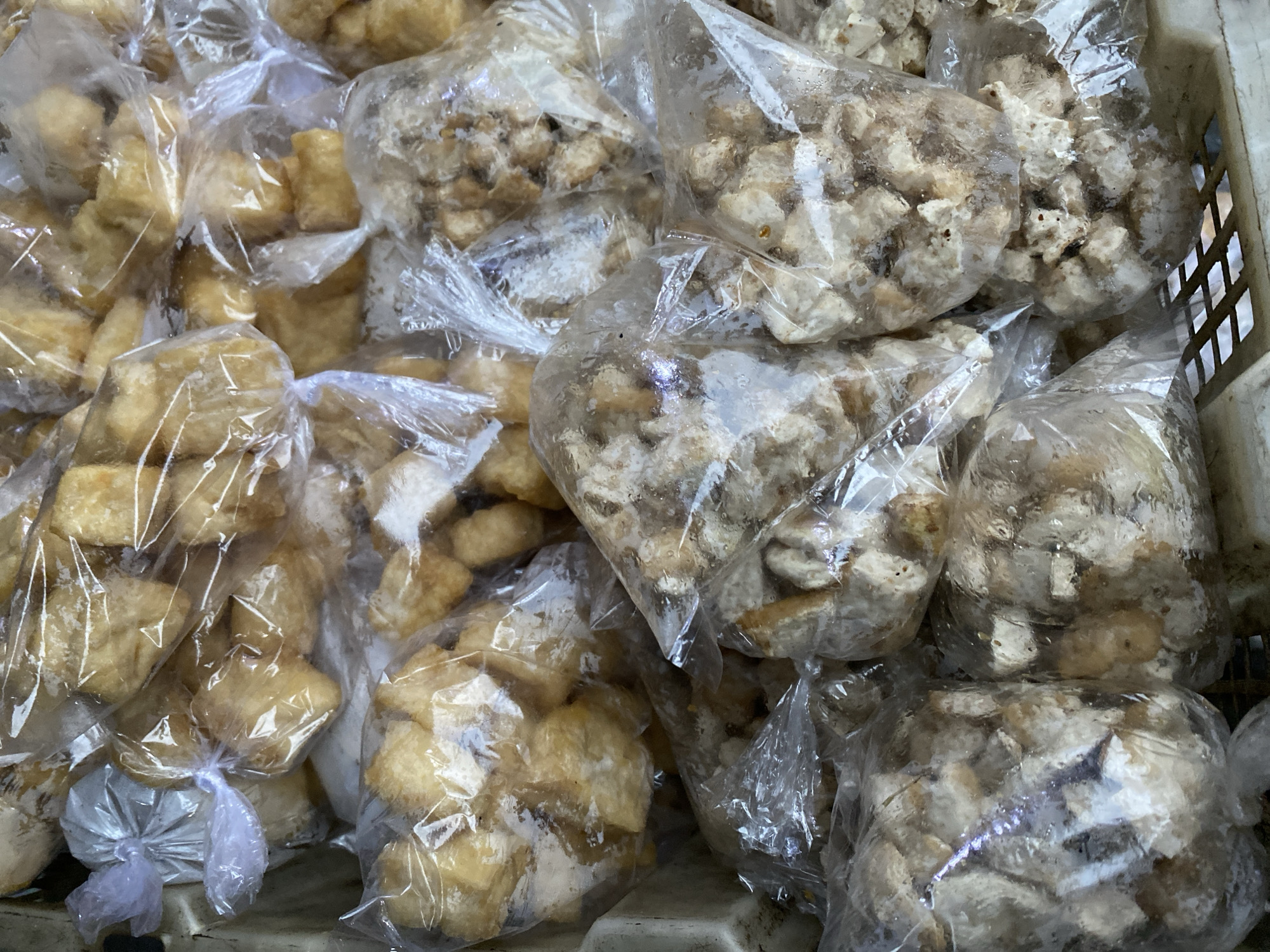 A close-up, overhead shot shows a large quantity of fried food items, possibly snacks or ingredients, packaged in clear plastic bags. Some bags are tied with white string. The food appears to be in bite-sized pieces, with some pieces having a golden-brown color, suggesting they are fried. Other pieces are lighter in color and appear more crumbly or irregular in shape. The bags are loosely filled and packed together in what appears to be a container or basket, some parts of which are visible at the edges of the frame, showing a textured, light-colored surface and a grid-like pattern at the bottom. The lighting is overhead and slightly diffused, casting some subtle shadows within the bags. The scene suggests a food vendor or market stall in Ambarketawang, Indonesia, where these items are prepared for sale.