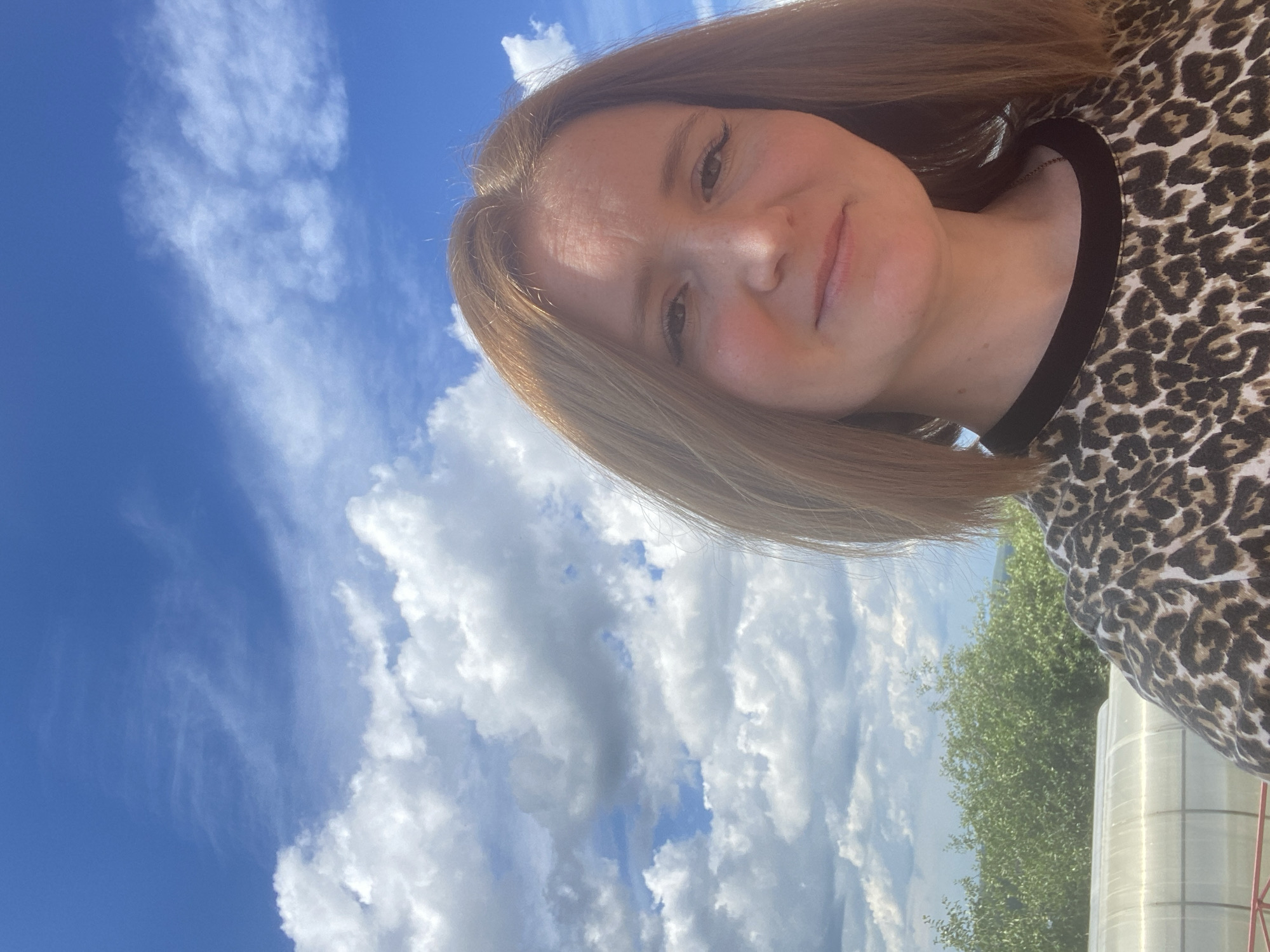 A young Caucasian woman with shoulder-length auburn hair and fair skin is pictured in a close-up selfie outdoors. She is wearing a black and brown leopard-print t-shirt with a black neckline. Her eyes are dark brown and she has dark eyeliner on. She is smiling faintly, with her head tilted slightly to the right. The background consists of a bright blue sky filled with fluffy white clouds. To the left and below the woman, there are green trees and what appears to be a greenhouse structure. The image is taken from a slightly upward angle. The lighting suggests it is daytime, likely a sunny day. The location is identified as Chaikovsky, Russia, given the context.