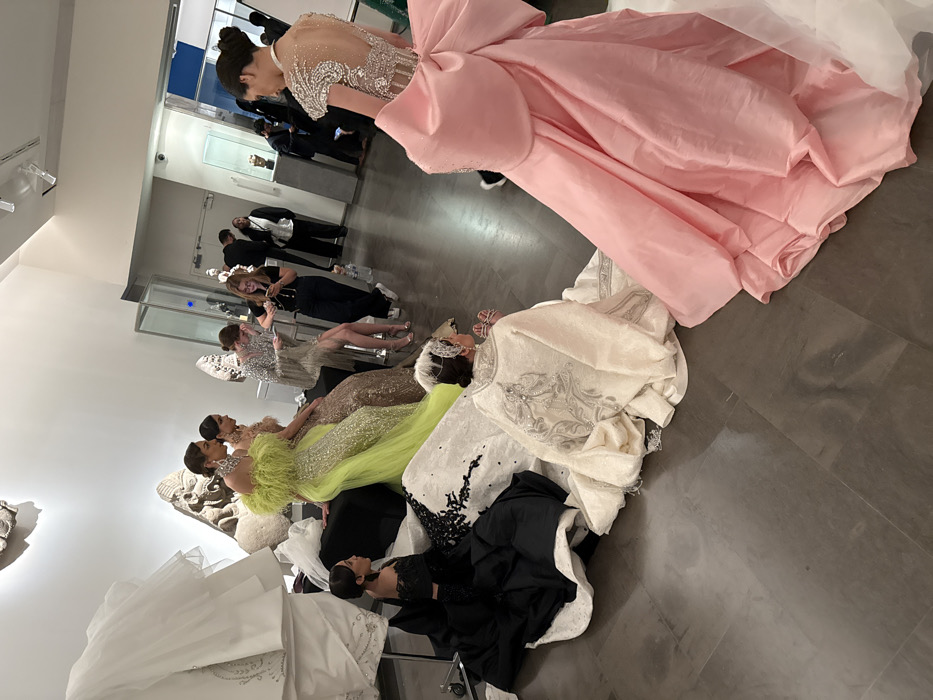 Several women, dressed in elaborate gowns of pink, black, green, tan, and white, are lined up in what appears to be a museum or gallery setting. The gowns are highly detailed with embellishments and dramatic silhouettes. A group of people in formal attire stand in the background behind a glass wall, with one individual taking a photo of the models. The setting suggests a fashion show or presentation.