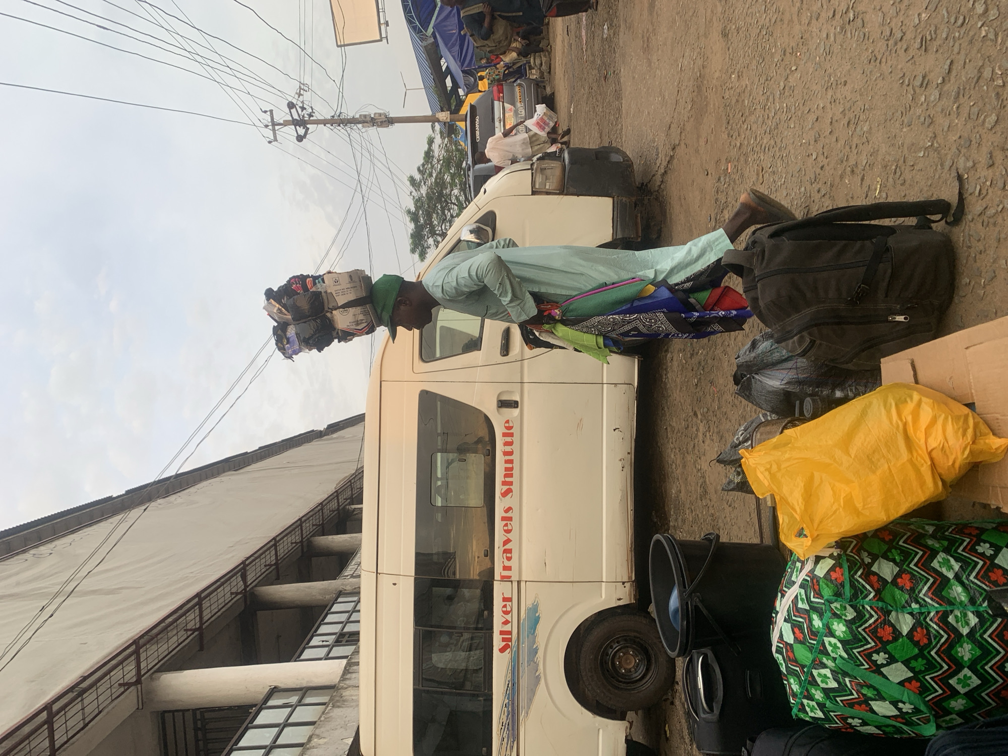 This is a medium shot, outdoor photograph taken during the day in Elelenwo, Nigeria. The scene depicts a man carrying a large, stacked load of bags and parcels on his head. He is wearing a light blue traditional outfit and a green cap. He is standing next to a white minibus with "Silver Travels Shuttle" written on its side. In the foreground, there are several bags and a black bucket on the ground. The background shows a building, electrical poles, wires, and other vehicles and market stalls. The ground is paved with rough asphalt. The sky is overcast.