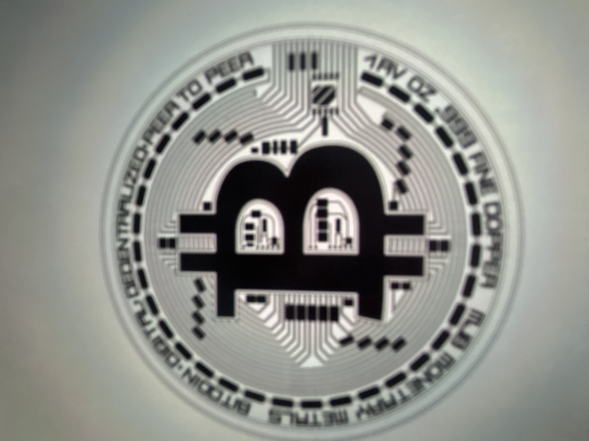 The image shows a close-up of a Bitcoin coin. The coin has the Bitcoin logo, a "B" inside a circle, on the front. Around the logo, text is visible which appears to read "BITCOIN DIGITAL DECENTRALIZED PEER TO PEER" and "METALS MONETARY 1AV OZ .999 FINE COPPER". The background is a blurry, light gray. There is no visible city or location information in the image.