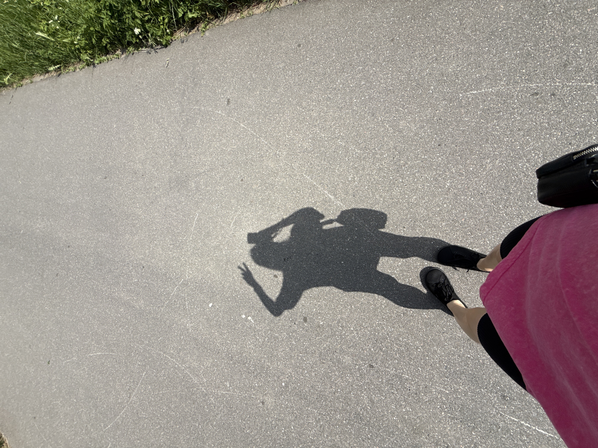 An over-the-shoulder, first-person perspective shot captures a shadow of a person on a paved asphalt path. The shadow, indicating a person holding a phone to take the photo, shows them with their left hand raised in a peace sign and their right hand holding a bag. The shadow's feet are clad in black sneakers, and the person appears to be wearing shorts with black leg coverings and a bright pink top. The path is bordered on the right by green grass and some small white flowers. The scene is lit by sunlight, casting a strong shadow. The location is a paved path in a park or along a street, likely in Saint Petersburg, Russia, given the lighting and vegetation.