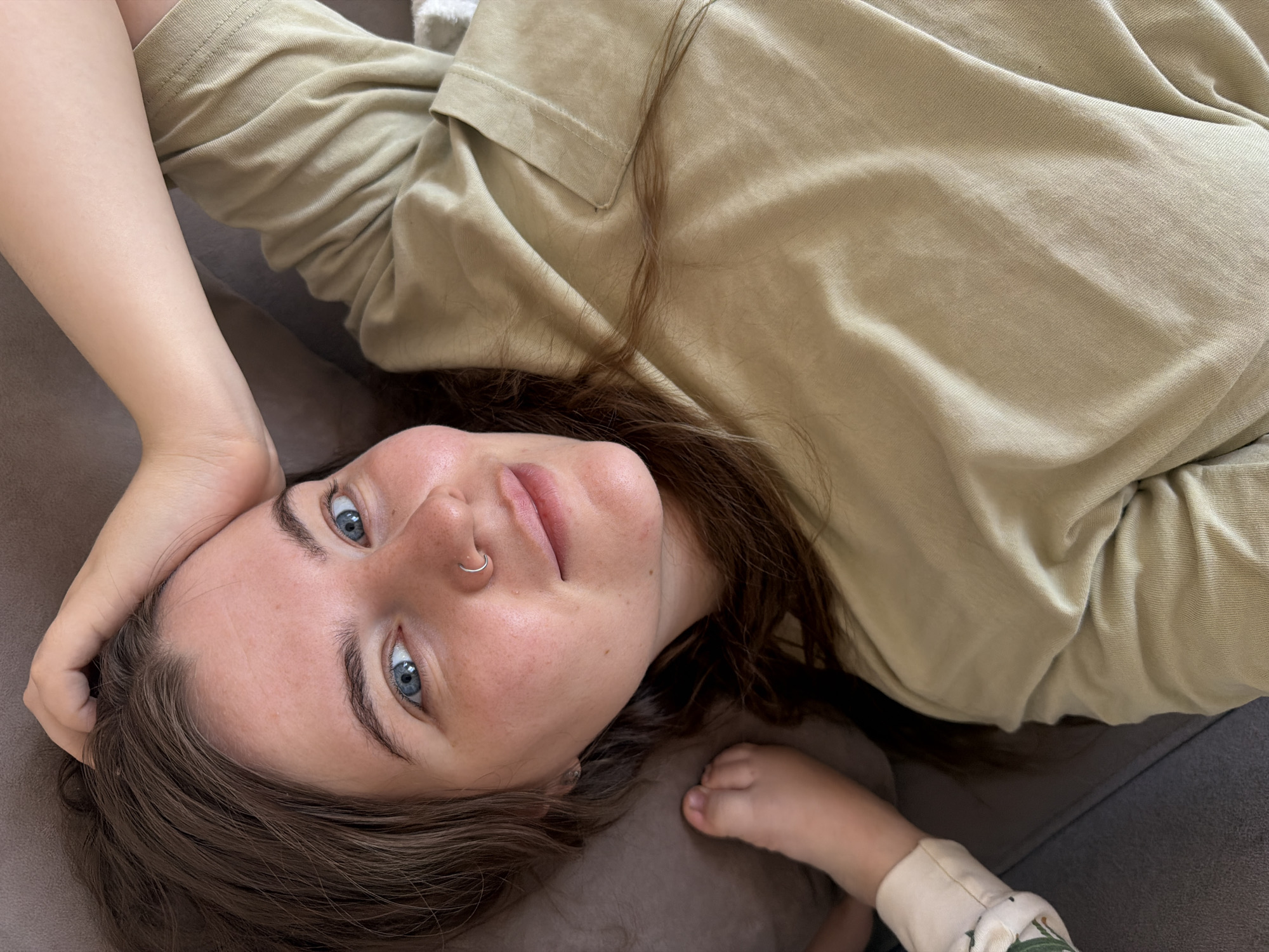 A woman is lying on a gray upholstered surface, likely a sofa. She is fair-skinned with brown hair and blue eyes and a silver ring in her left nostril. She is wearing a light-brown shirt with a collar. Her left hand is up near her head. The partially visible hand of an infant wearing a light-colored sleeve with a floral pattern is on the lower right. The image was taken in Chaikovsky, Russia.