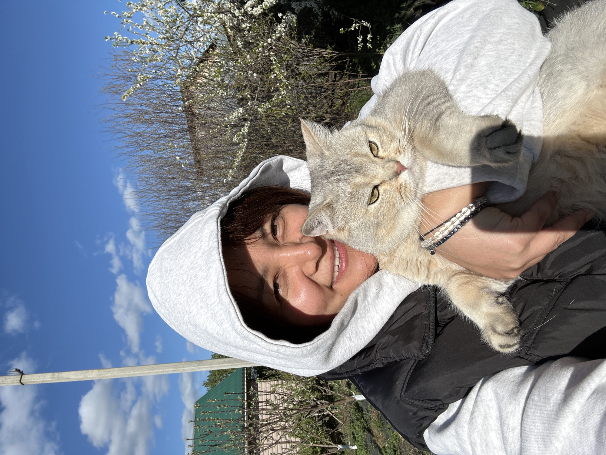 A woman in a white hoodie and black jacket is holding a light gray cat. The woman is smiling and looking at the camera, while the cat is looking slightly to the side. A white flowering tree, a pole, a house with a green roof, and a blue sky with light clouds are visible in the background. This photo may have been taken near Abzakovo, Russia.