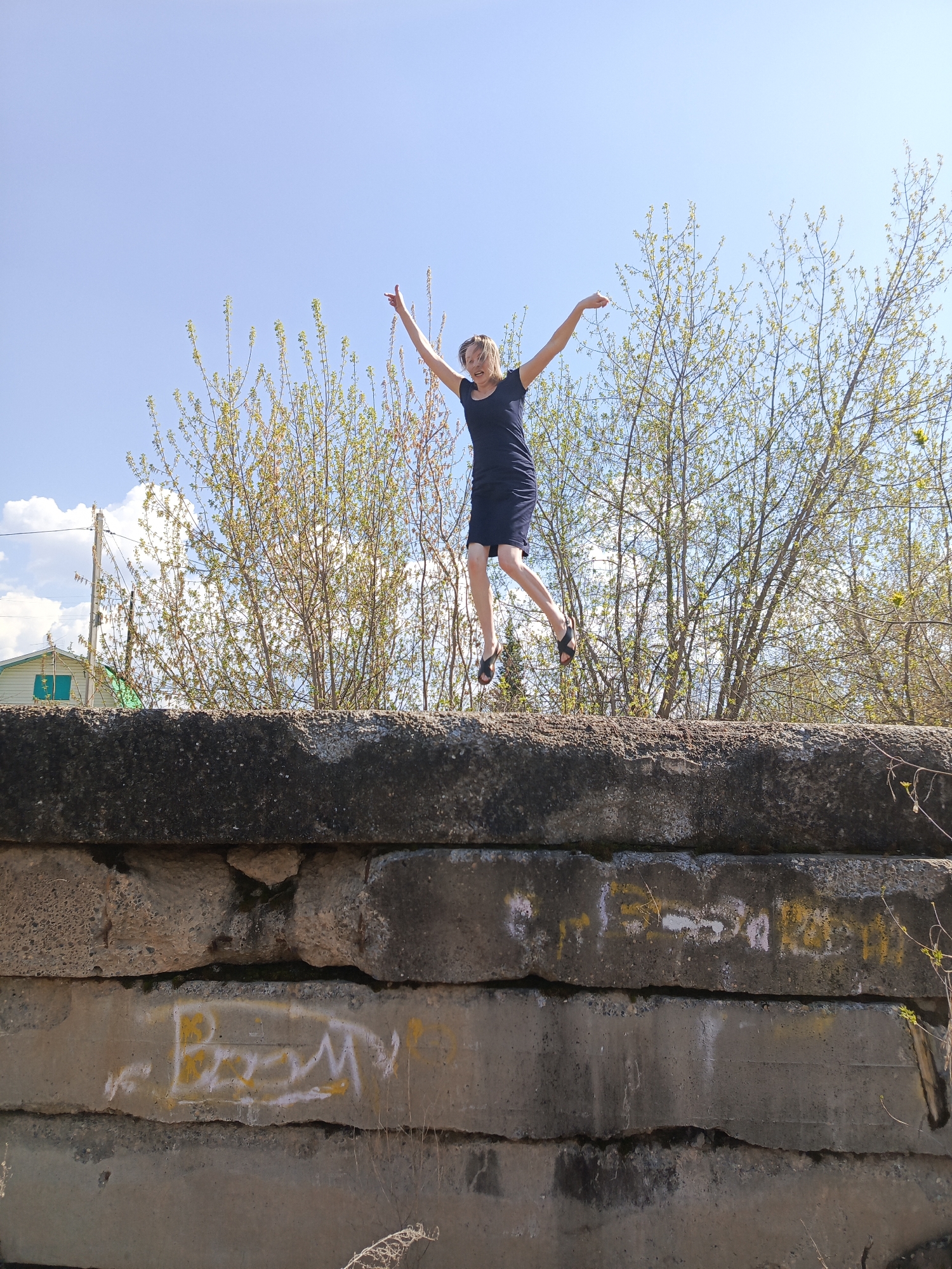 A young woman is captured mid-air, jumping on a concrete structure in Chaikovsky, Russia. She is wearing a dark-colored dress and sandals. Her arms are extended upwards, and she appears to be smiling. Behind her is a row of trees with emerging leaves, and a clear blue sky with some white clouds. To the left, a glimpse of a building with green trim and a utility pole is visible. The concrete structure is composed of large, stacked blocks with graffiti and weathering visible on the surfaces. The area in front of the structure has sparse vegetation.