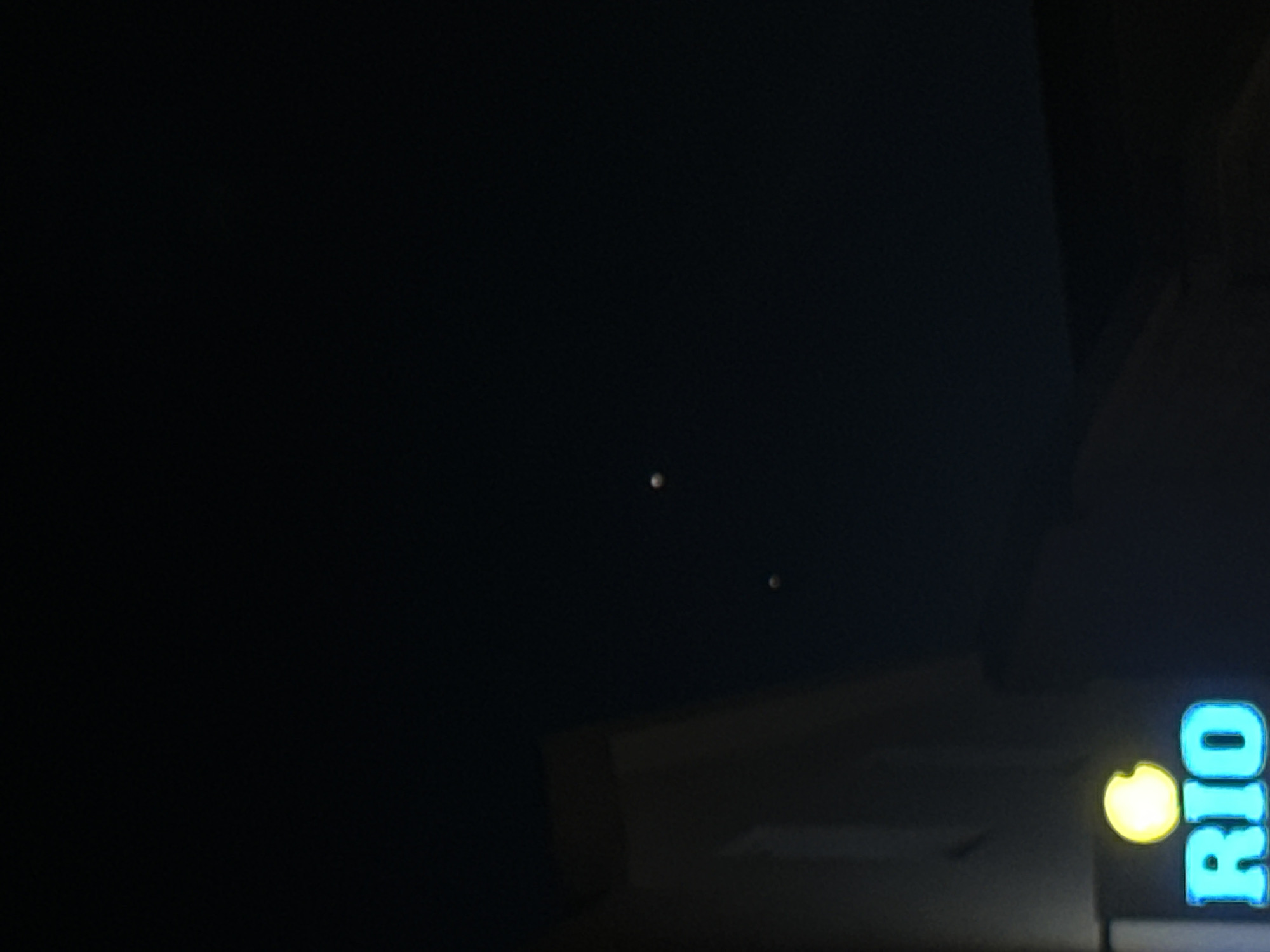 The image captures a nighttime scene in Budva, Montenegro. In the foreground, a brightly lit sign displays the word "RIO" in cyan letters, with a yellow circle above it. To the left of the sign, a portion of a building is visible, appearing as a light-colored structure against the dark sky. In the upper right quadrant of the image, two luminous, spherical objects are suspended in the sky.