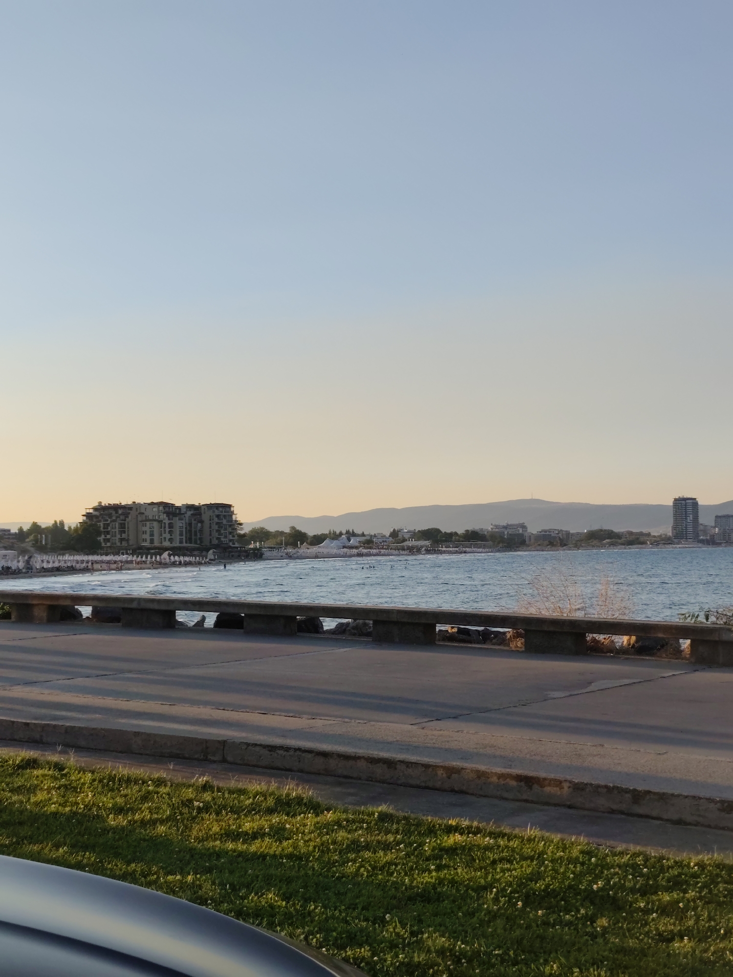 This image depicts a coastal scene in Nesebar, Bulgaria, likely during the late afternoon or early evening, given the warm, soft lighting. In the foreground, a paved walkway runs parallel to the sea, bordered by a low concrete barrier with stone supports. Beyond the barrier, the blue sea stretches towards the horizon, with gentle waves lapping against a sandy beach.
Along the beachfront, a line of white beach umbrellas suggests a public area, with scattered individuals visible enjoying the water and the shore. Several buildings are visible in the mid-ground, including a large, multi-story hotel or apartment complex with a tan facade, situated behind the beach. Further down the coastline, other buildings and structures, including a tall, slender skyscraper, punctuate the distant landscape, which is backed by rolling hills.
The sky above is a clear gradient of light blue, transitioning to a pale yellow or cream hue near the horizon, indicating the sun is low in the sky. Green grass borders the paved area in the immediate foreground, with shadows from the concrete barrier stretching across the walkway. A portion of a dark-colored vehicle is visible in the bottom left corner, suggesting the perspective is from a street or parking area adjacent to the promenade. The scene captures a serene moment of a popular seaside resort town.