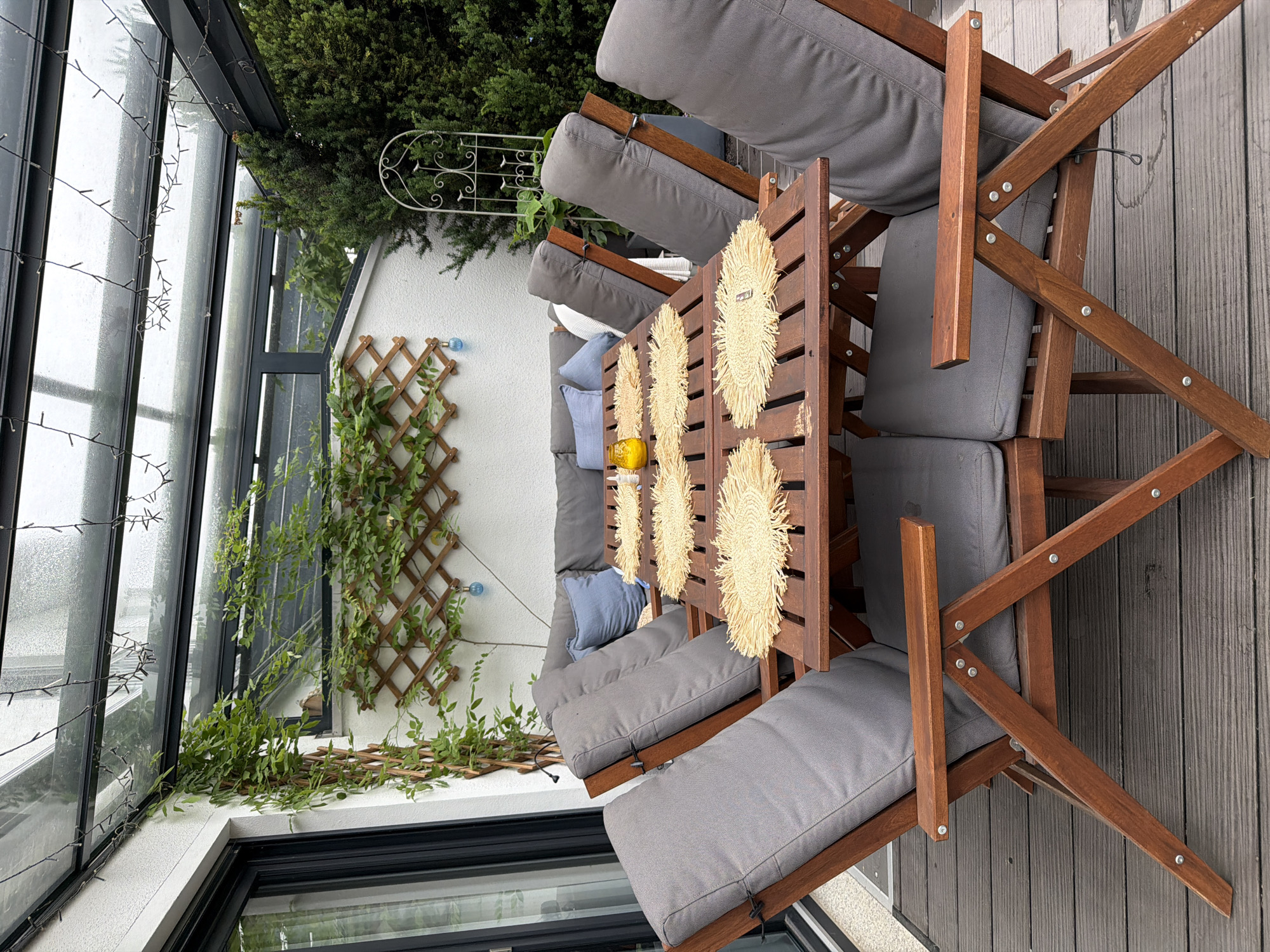 This image depicts an outdoor seating area with a wooden table and chairs, likely in Poing, Germany. The furniture is arranged on a dark wooden deck beneath a glass-covered roof structure, which is strung with decorative lights. The table is set with woven placemats and a small yellow glass candle holder. Several grey cushioned chairs are positioned around the table. In the background, a white wall is adorned with a wooden trellis, overgrown with green vines. Beyond the trellis, a dense green hedge and a white decorative metal garden fixture are visible. The overall scene suggests a comfortable, contemporary outdoor living space.